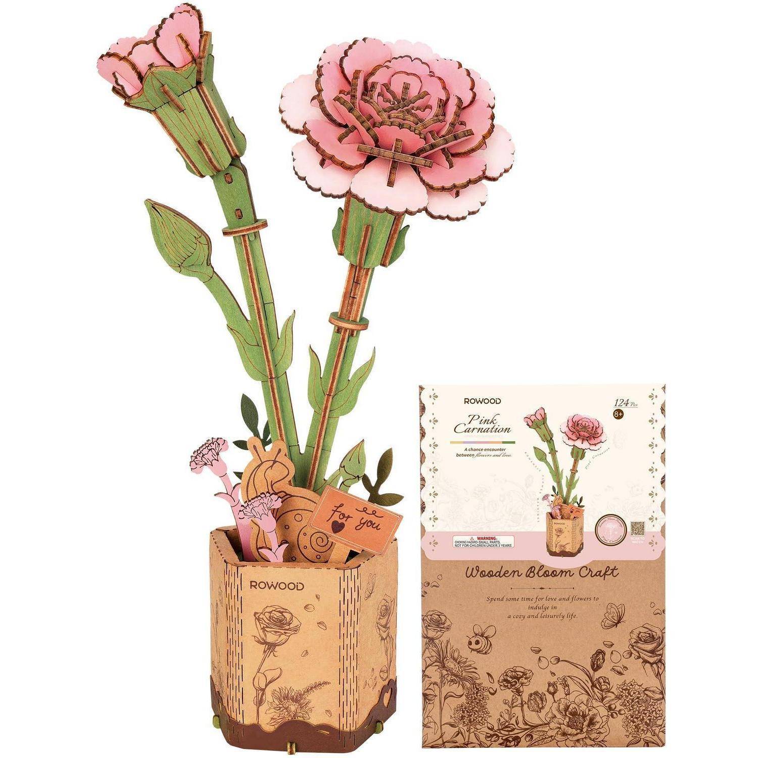 Ein Holz-3D-Nelken-Bastelset, das montiert mit rosa Blüten in einem Blumenkübel gezeigt wird, daneben seine Produktschachtel mit der Aufschrift 