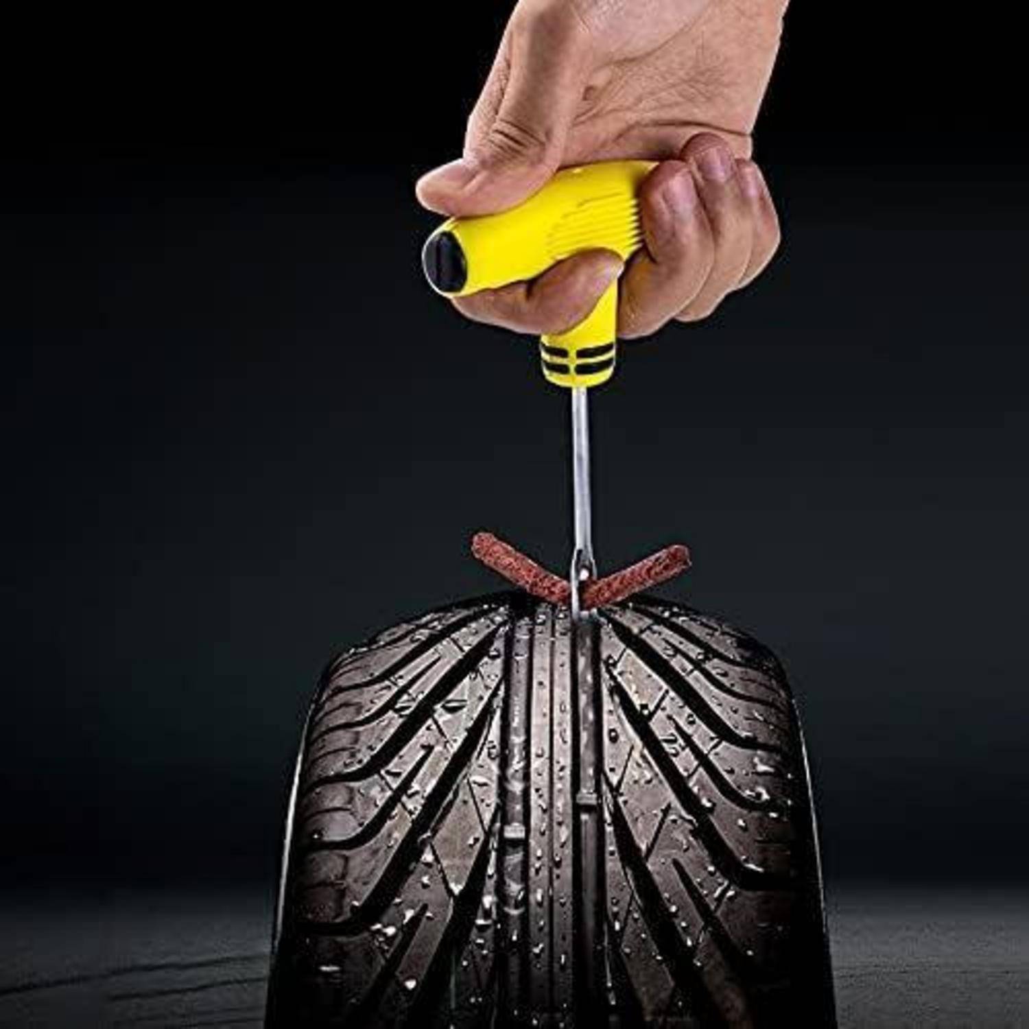 Hand hält einen Schraubendreher, der in die Lauffläche eines Autoreifens eingeführt ist, wobeieine Perforation oder Reifenreparatur an einem nassen Reifen vor dunklem Hintergrund demonstriert wird.