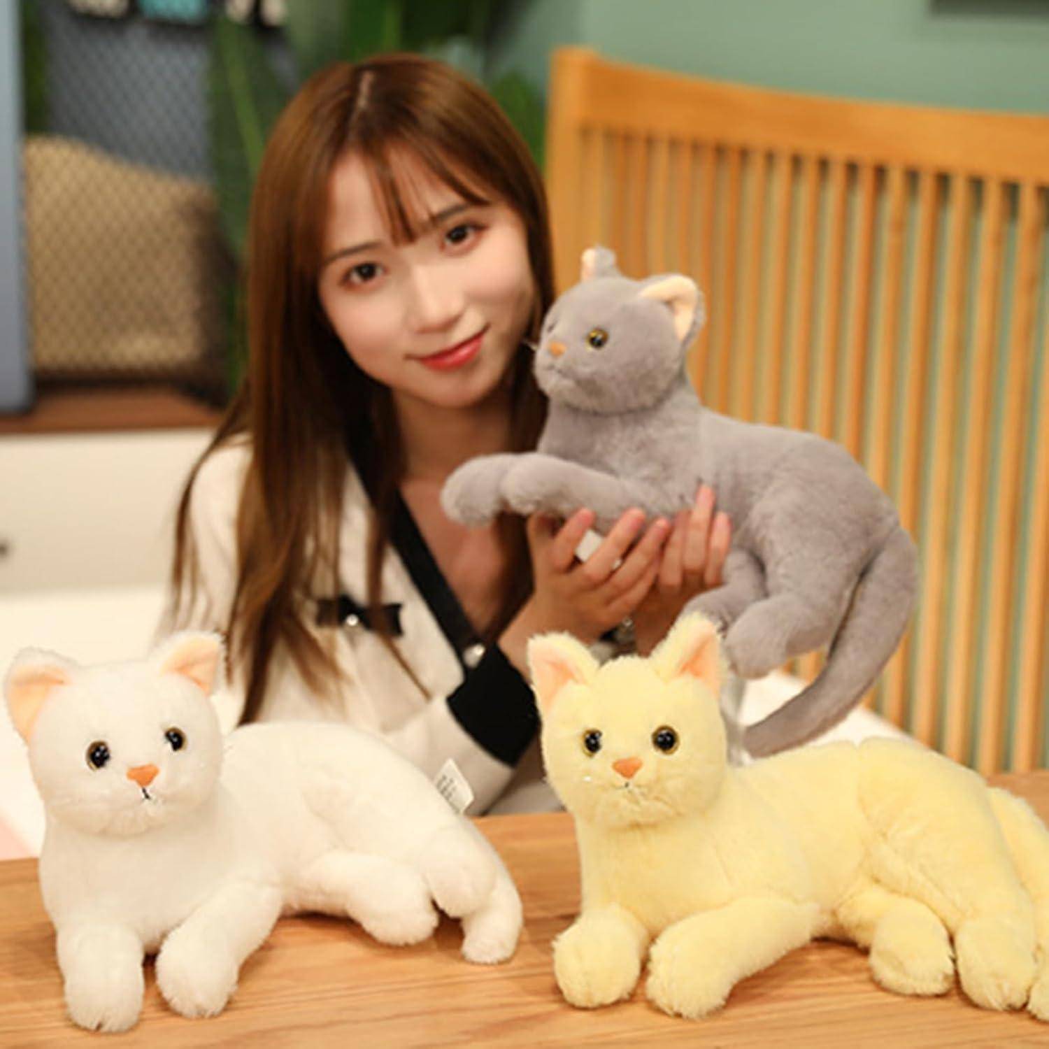 Junge Frau, die eine graue Plüschkatze hält, mit weißen und gelben Kuscheltieren, die auf einem Tisch in einer gemütlichen Inneneinrichtung ausgestellt sind.