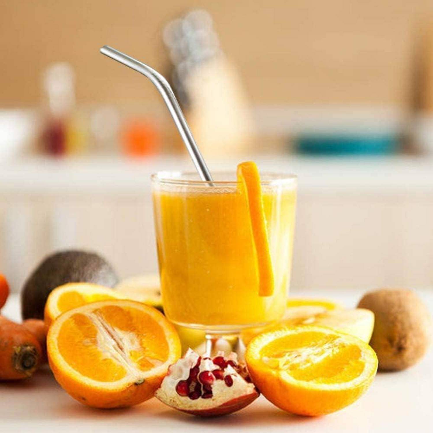 Glas mit orangefarbenem Smoothie mit einem Metallstrohhalm, umgeben von geschnittenen Orangen, Granatapfel, Avocado und Kiwi auf einer Küchentheke.