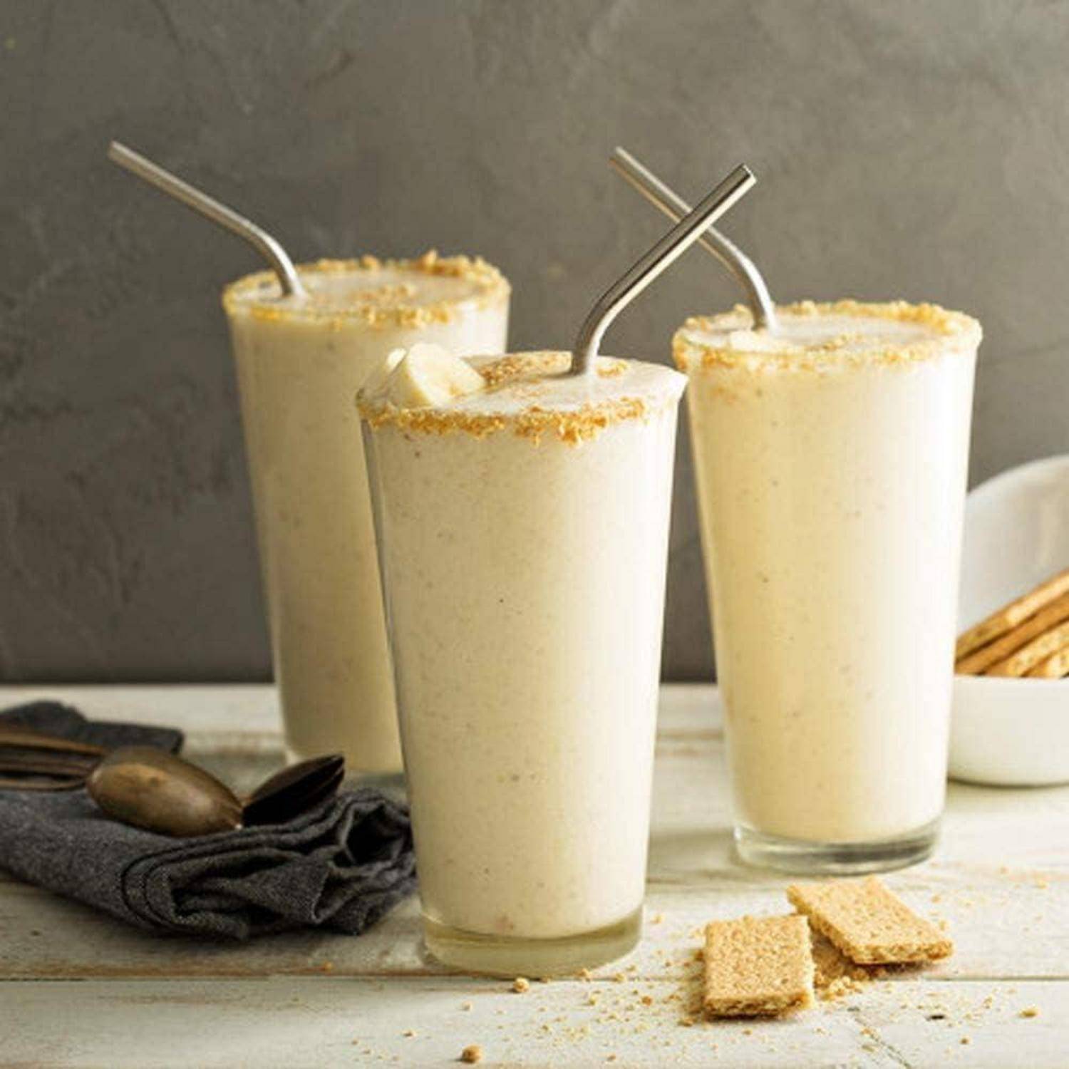 Drei schaumige, helle Smoothies oder Milchshakes in durchsichtigen Gläsern mit Metallstrohhalmen, oben mit Krümeln bestreut, auf einem Tablett mit Servietten und Keksstücken.