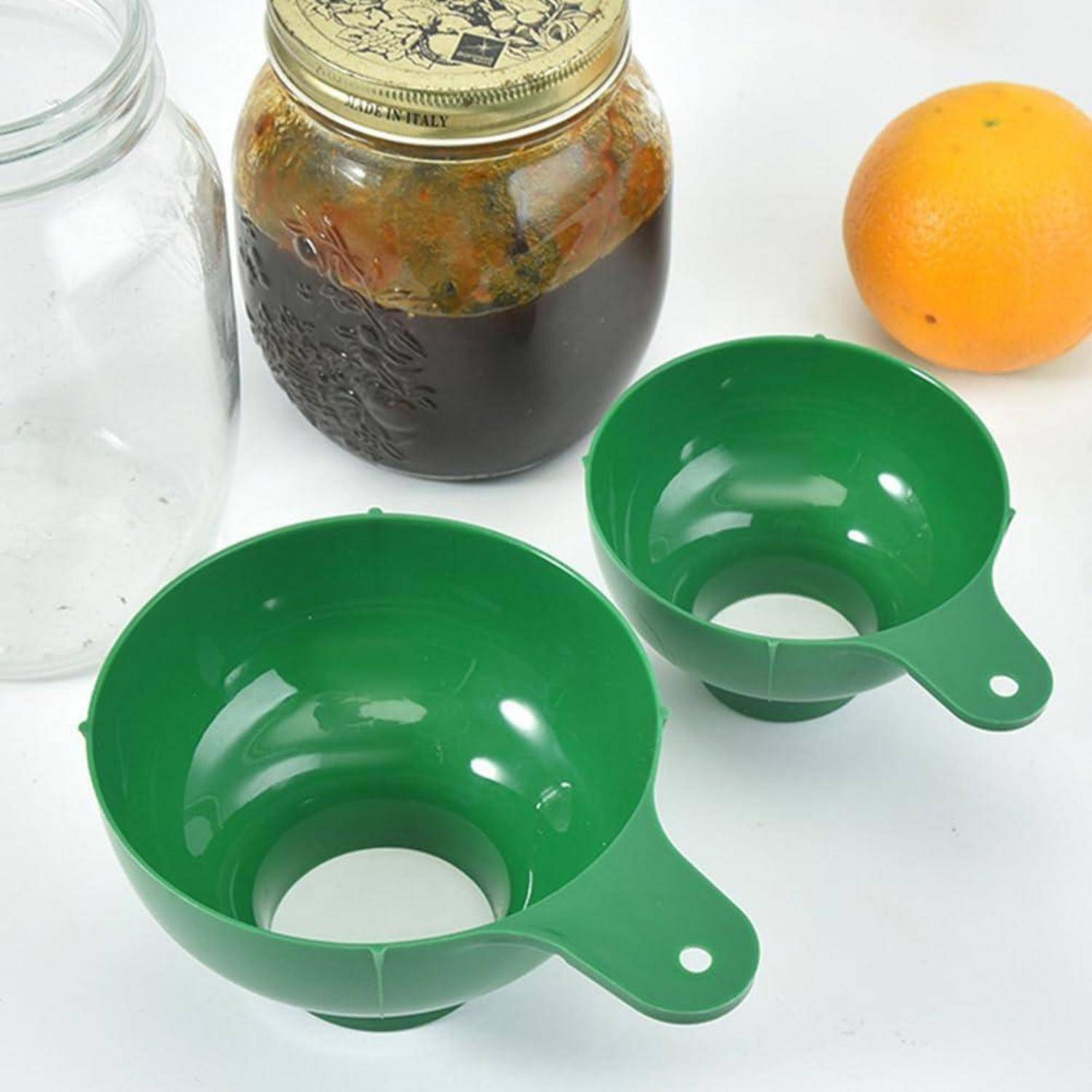 Zwei grüne Kunststofftrichterbecher vor einem Glas, ein leeres Glas und eine Orange auf einer weißen Oberfläche.