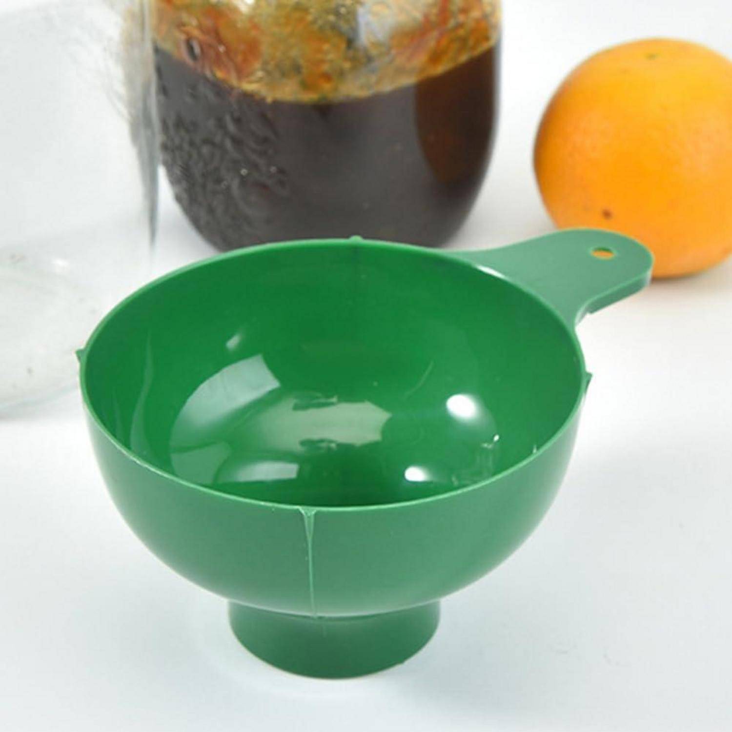 Grüne Kunststofftrichter-Schüssel mit Griff, gezeigt auf einer weißen Oberfläche mit einem Glas und einer Orange im Hintergrund.