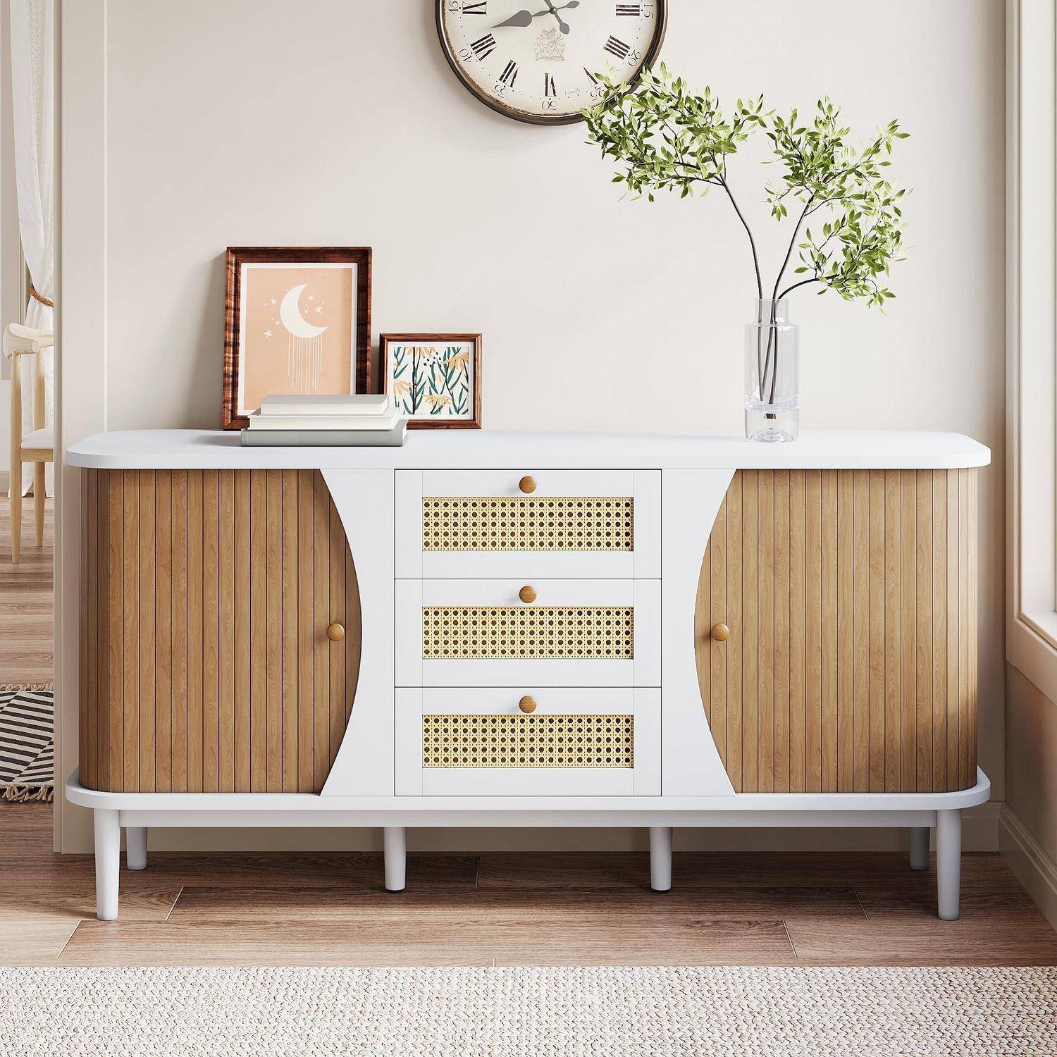 Weißes Sideboard mit Holzschranktüren und drei Schubladen mit Korbgeflecht-Front, oben dekoriert mit gerahmter Kunst, Büchern und einer Vase mit Grünpflanzen unter einer Wanduhr.