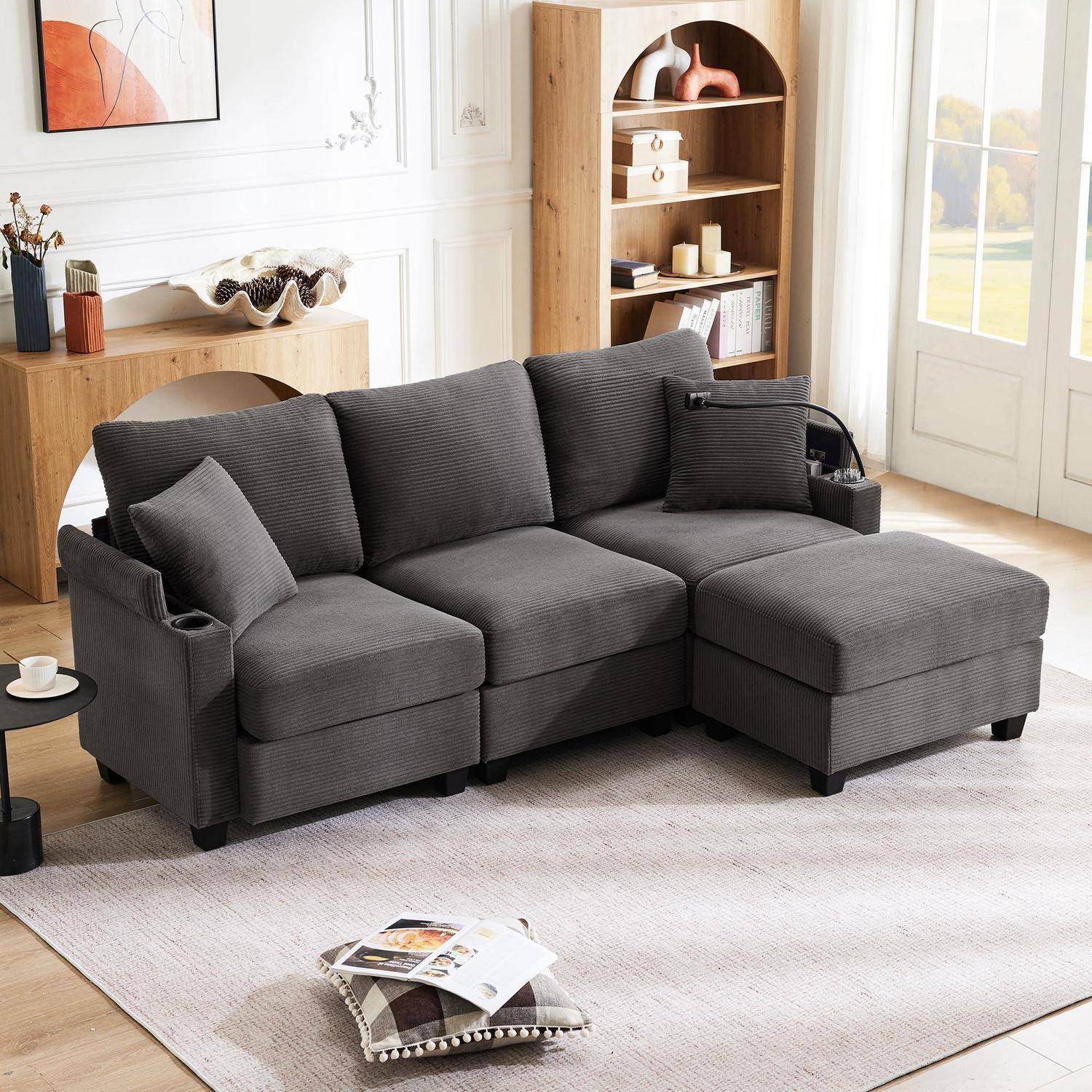 Graues L-förmiges Ecksofa mit Chaiselongue in einem hellen Wohnzimmer, mit Beistelltisch, Bücherregal, Teppich und Zeitschriften auf dem Boden.