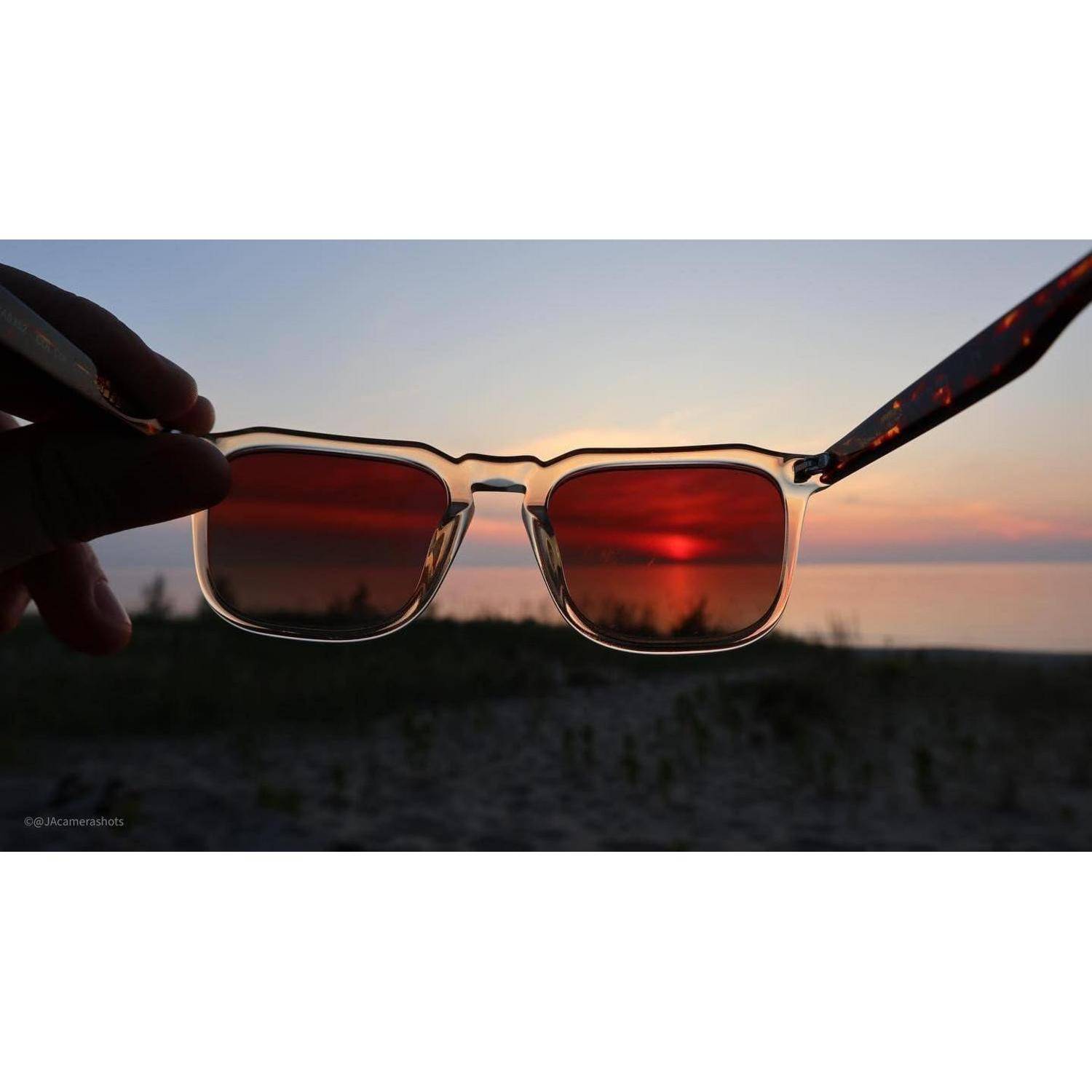 Hand hält Sonnenbrille mit rotgefärbten Gläsern vor einem Sonnenuntergang über dem Wasser, mit der Sonne gespiegelt in den Gläsern.
