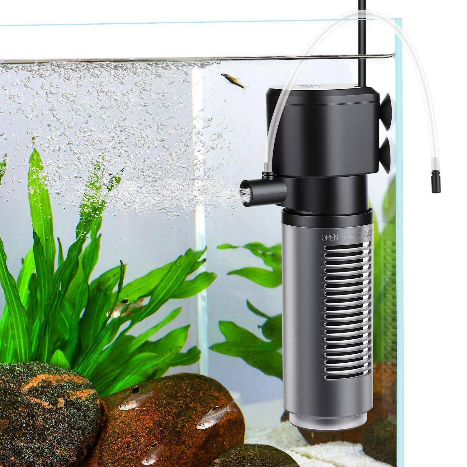 Aquariumecke mit schwarzem internem Filter und transparentem Schlauch, neben grünen Pflanzen, Steinen und kleinen Fischen.
