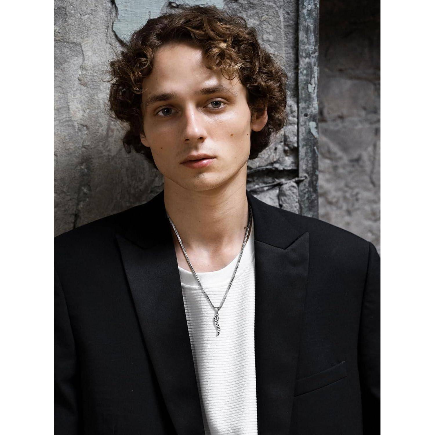 Porträt eines jungen Mannes mit lockigem Haar, der einen schwarzen Blazer über einem weißen Hemd und einer Halskette trägt und gegen eine rauhe Steinmauer lehnt.