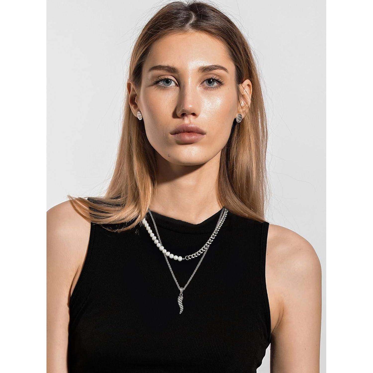 Porträt einer jungen Frau mit gllattem blondem Haar, das ein schwarzes ärmelloses Top, eine Perlenkette und Ohrringe gegen einen einfachen hellen Hintergrund trägt.