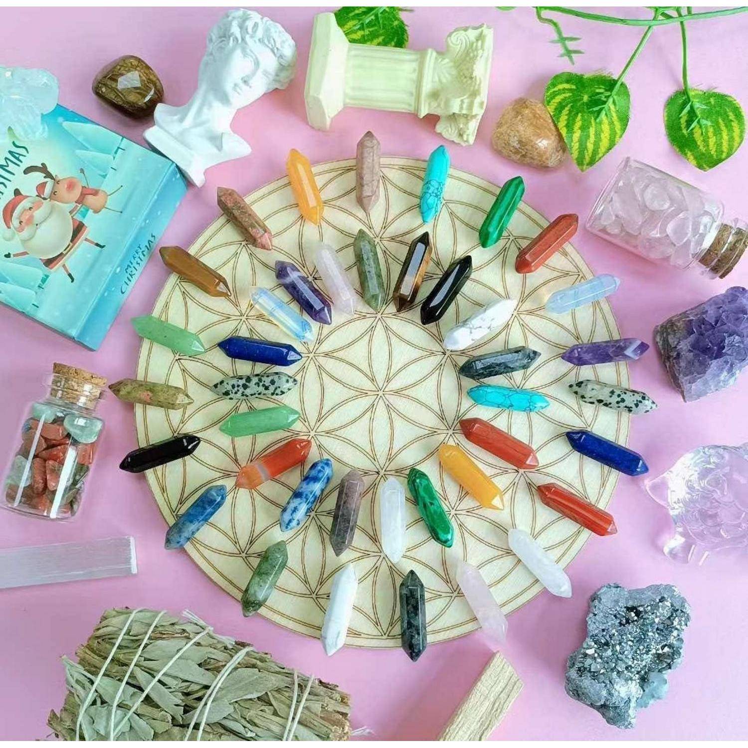 Kristallheilungs-Display mit farbenfrohen polierten Steinen, die in einem Kreis auf einem geometrischen Mandala-Brett angeordnet sind, umgeben von Kräutern, kleinen Flaschen und Mineralienproben auf rosa Hintergrund.