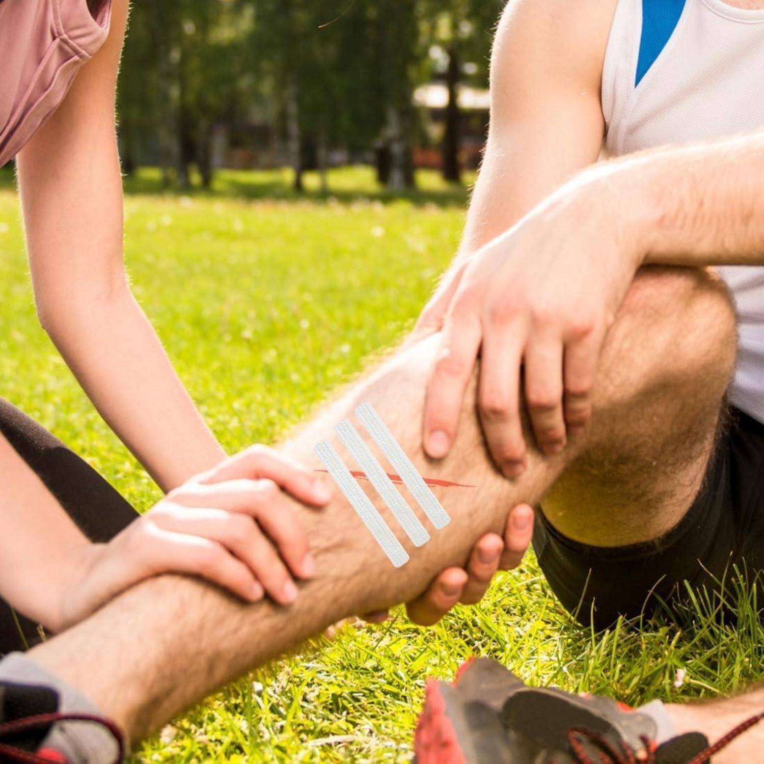 Eine Person, die einem verletzten Mann im Freien in einem grasbewachsenen Park Verbandsmaterial um das Bein anlegt.