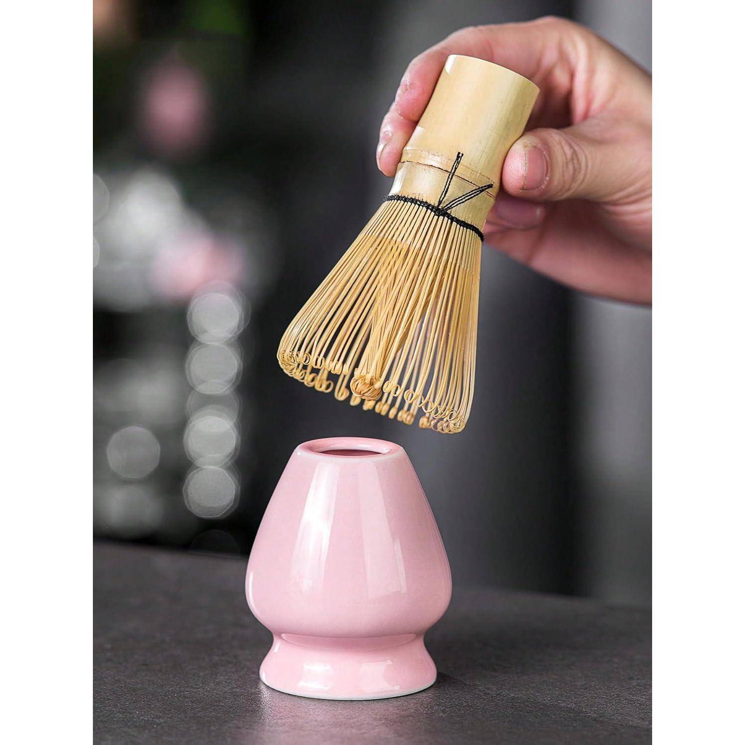 Hand senkt einen Bambus-Matcha-Schneebesen in einen rosa Keramikhalter auf einer dunklen Arbeitsplatte.