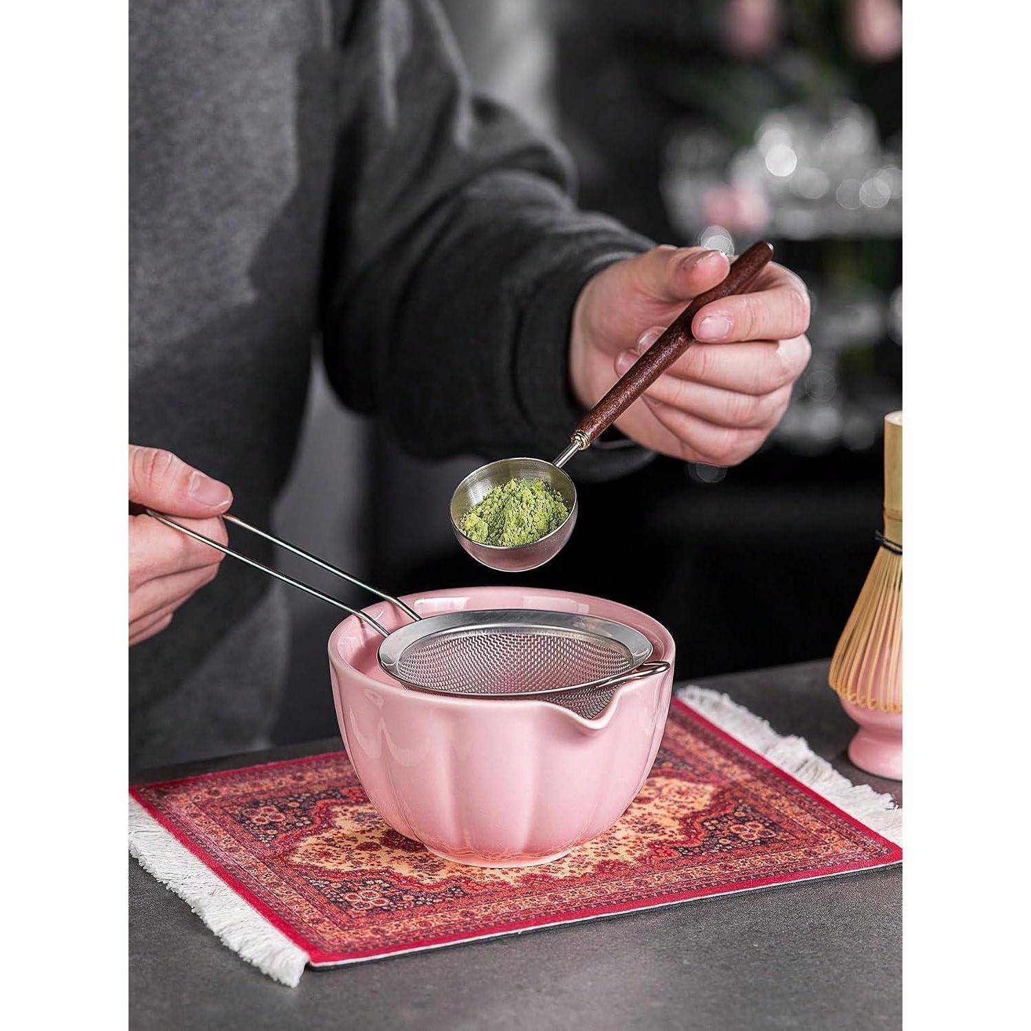 Person bereitet Matcha-Tee zu, schöpft grünes Pulver mit einem Sieb in eine rosa Schüssel auf einer gemusterten Matte.