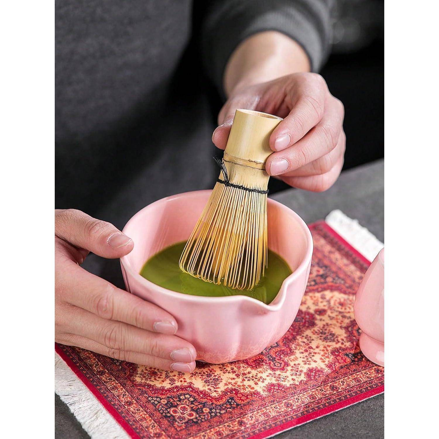 Hände, die grünes Matcha in einer rosa Schüssel mit einem Bambus-Chasen auf einem gemusterten Tuch aufschlagen.