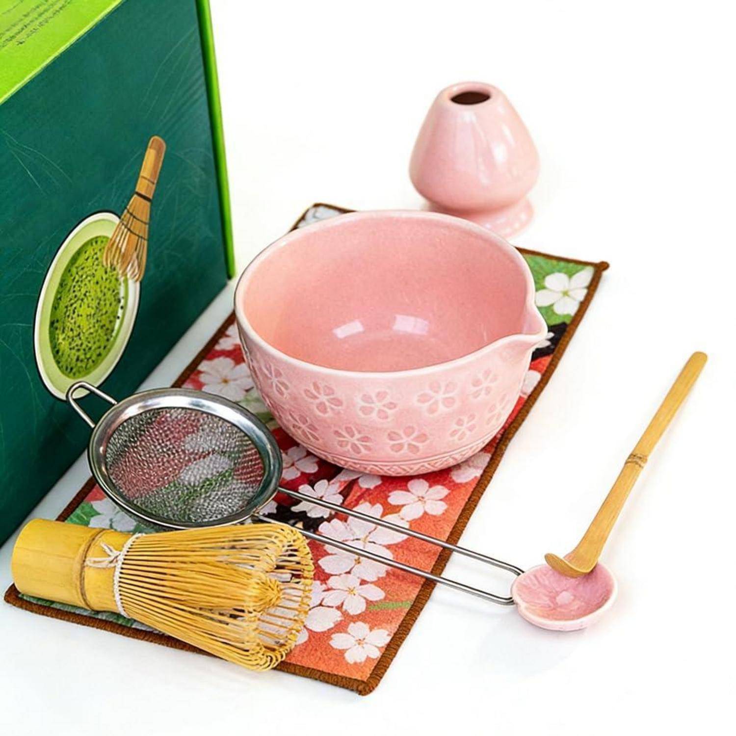 Rosa Matcha-Tee-Set mit Schale, Besen, Löffel, Sieb und Behälter auf einer Blumenmatte.