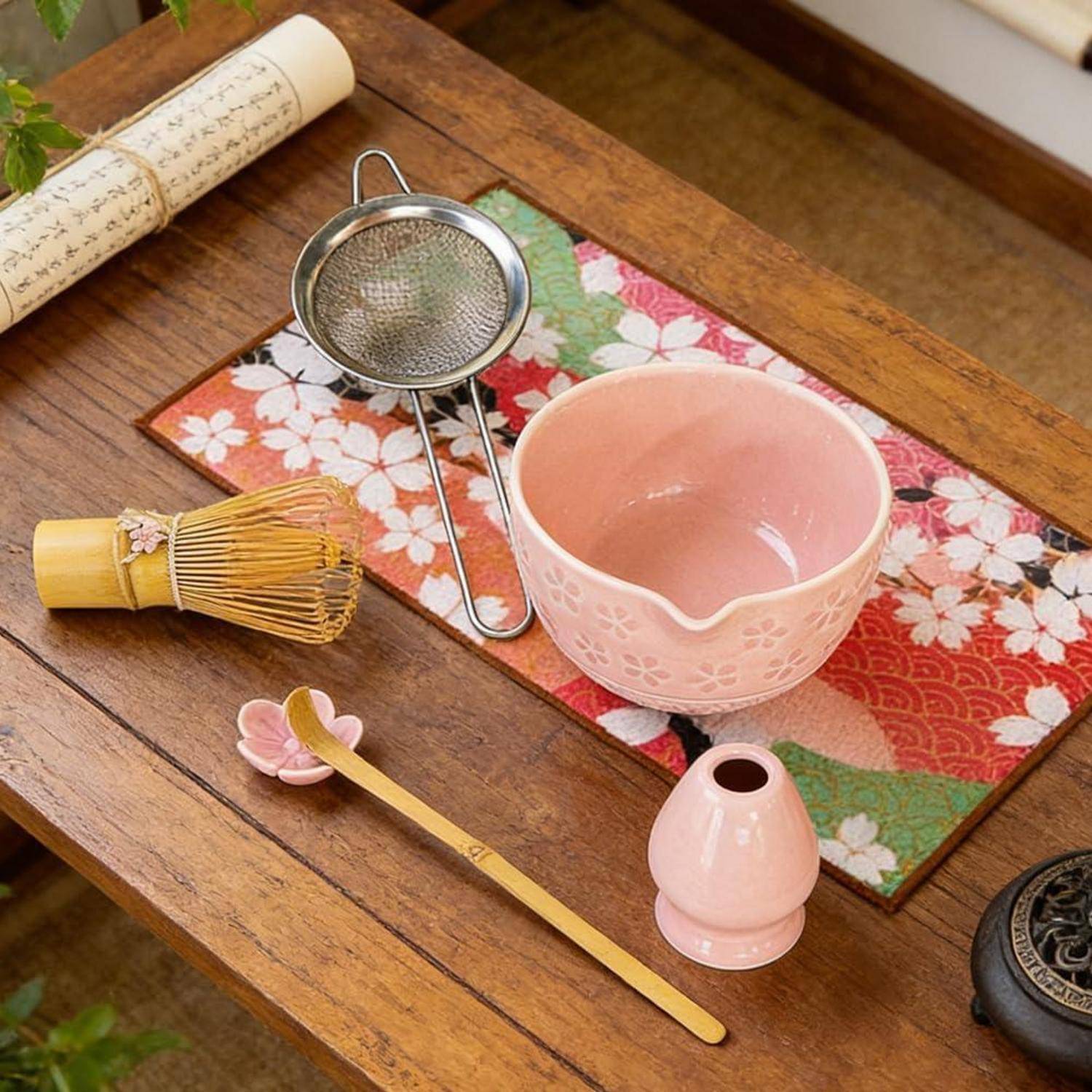 Teezeremonieset auf einem Holztisch, mit einer rosa Schale, Bambusbesenquaste, Sieb, Löffel und gemusterter Tischdecke mit Blumenmotiven.