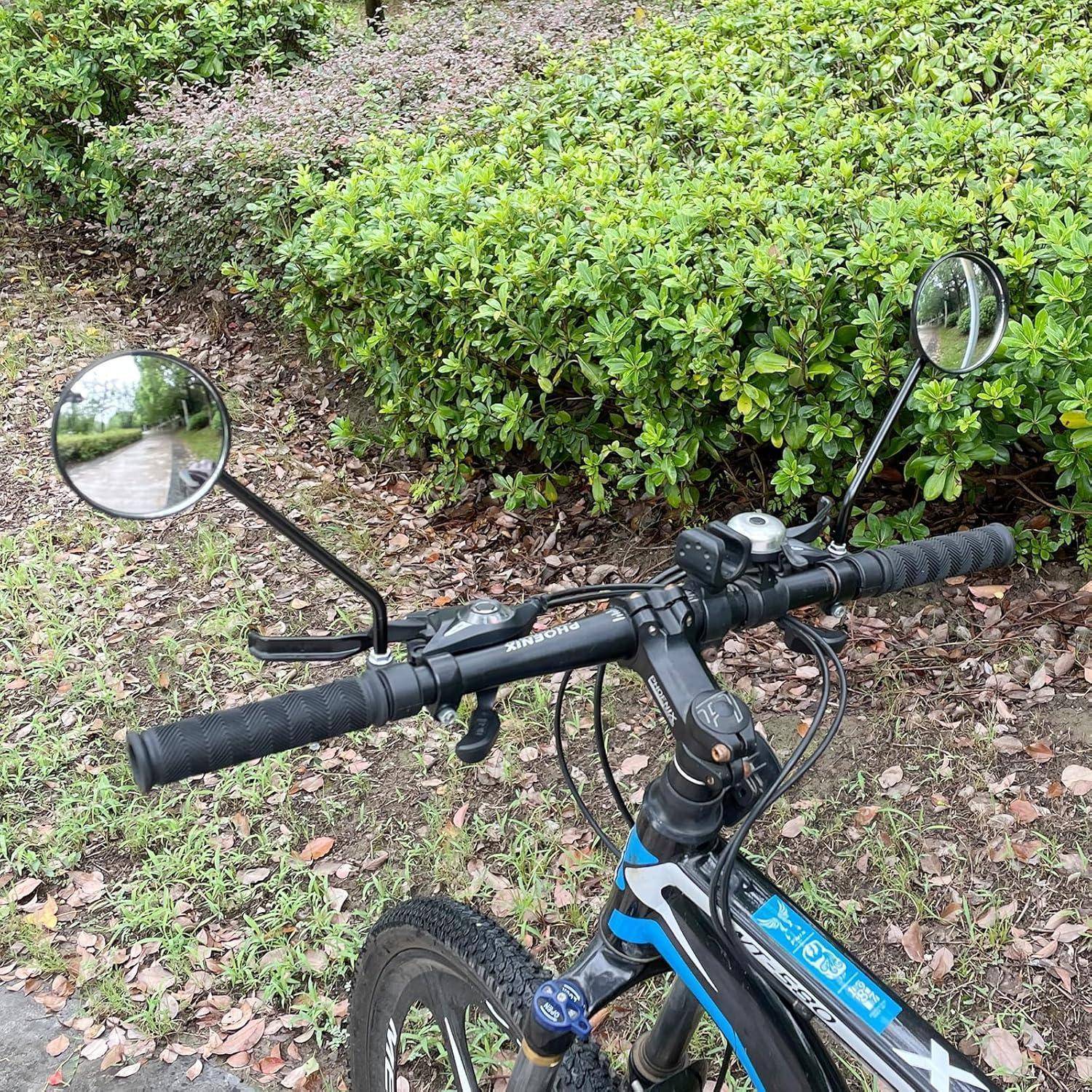 Nahaufnahme eines Fahrradlenkers mit zwei Rückspiegeln, geparkt neben einer Hecke auf einem blättrigen Weg.