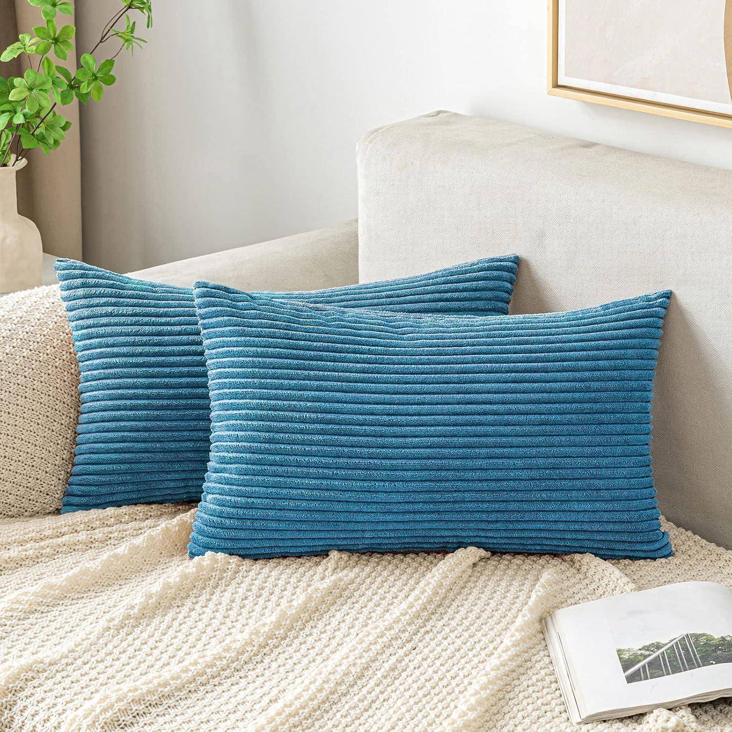 Zwei blaue gerippte rechteckige Wurfkissen auf einem beige Sofa mit einer cremeweißen Strickdecke und einem aufgeschlagenen Buch in einem hellen Wohnzimmer.