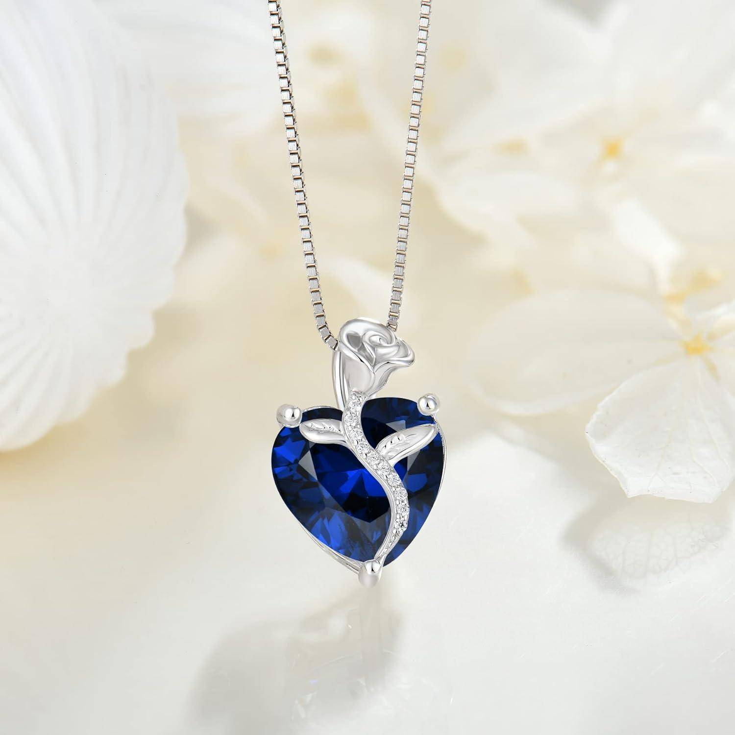 Silberner Anhängernecklace mit einem tiefblauen herzförmigen Edelstein, akzentuiert durch eine zarte Ranke und kleine klare Steine, auf einem weichen floralen Hintergrund.