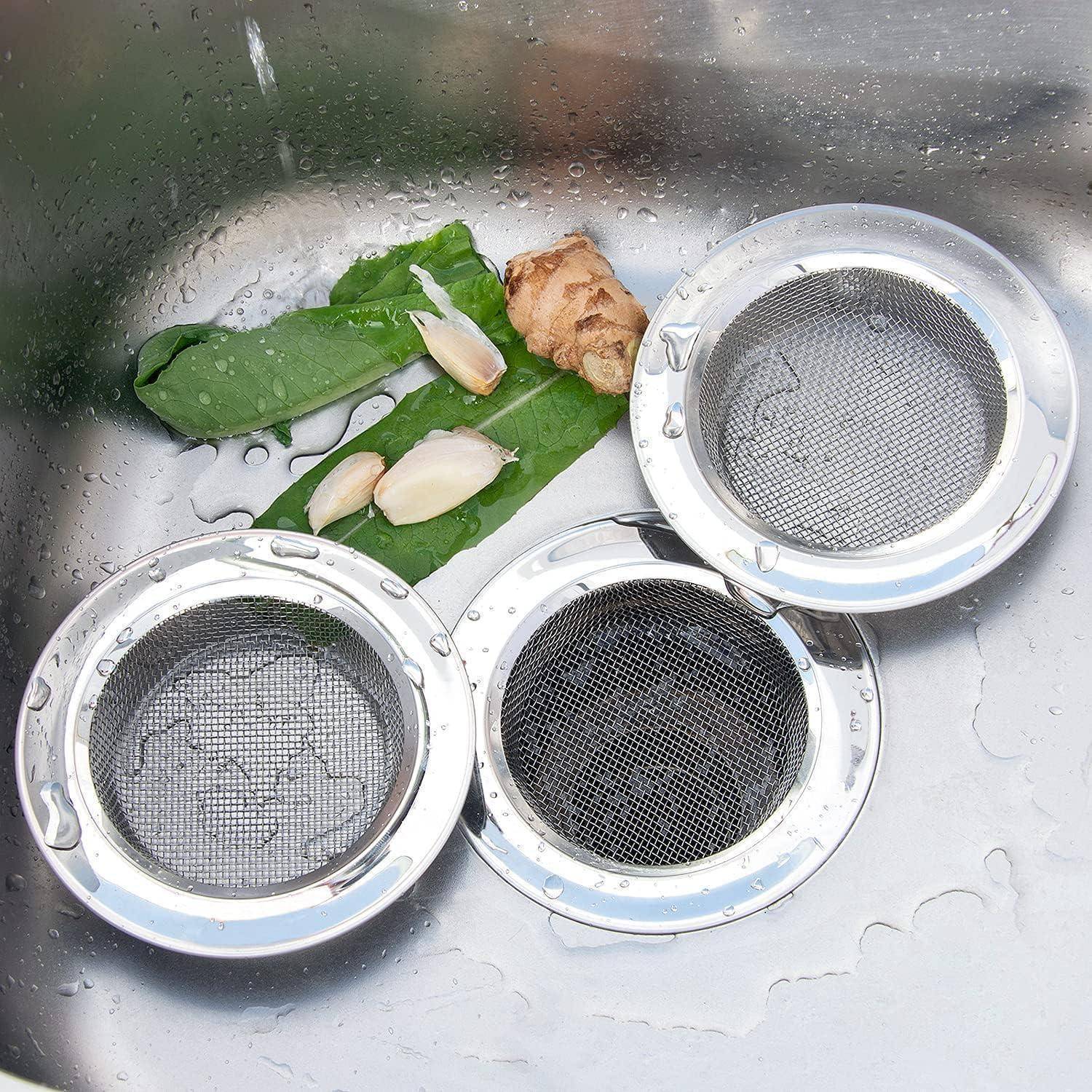 Drei Metallspülensiebe in einem Edelstahlspülbecken mit Wasser, daneben grüne Blätter, Knoblauchzehen und ein Stück Ingwer.