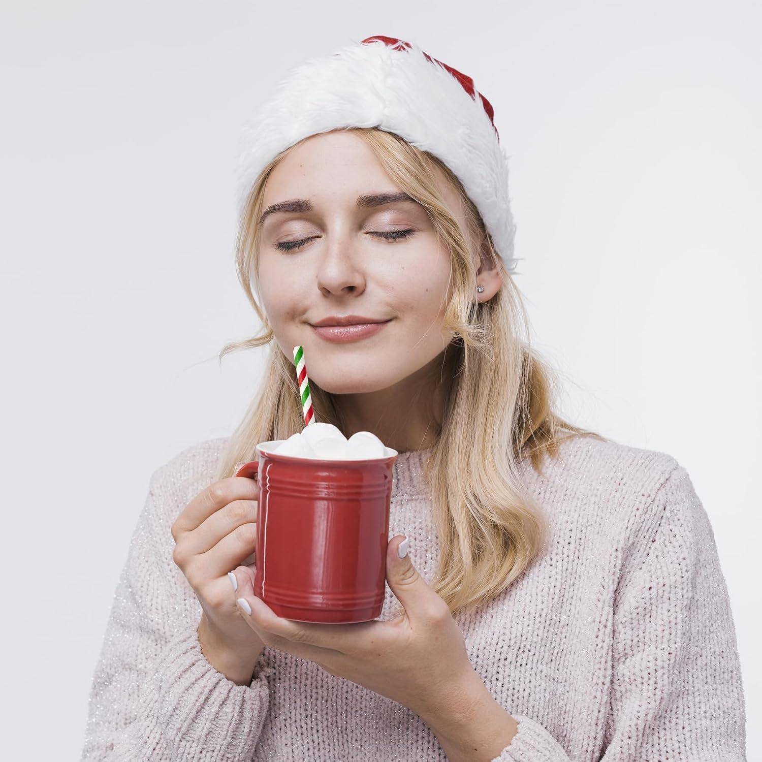 Lächelnde Frau in einer Weihnachtsmütze hält einen roten Becher mit heißer Schokolade, der mit Schlagsahne und einem gestreiften Zuckerstangenstab garniert ist.