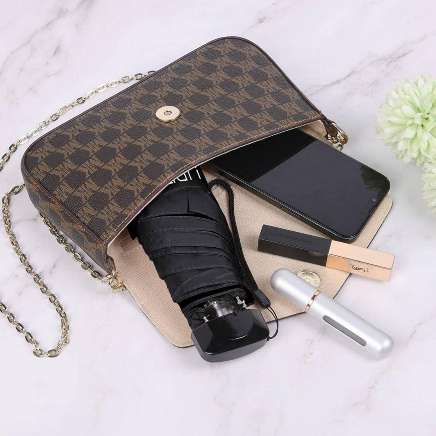 Offene braune Monogramm-Schultertasche mit Kettengurt, darin ein zusammengefalteter Regenschirm, ein Smartphone, Kosmetikprodukte und eine kleine Taschenlampe auf einer hellen Oberfläche.