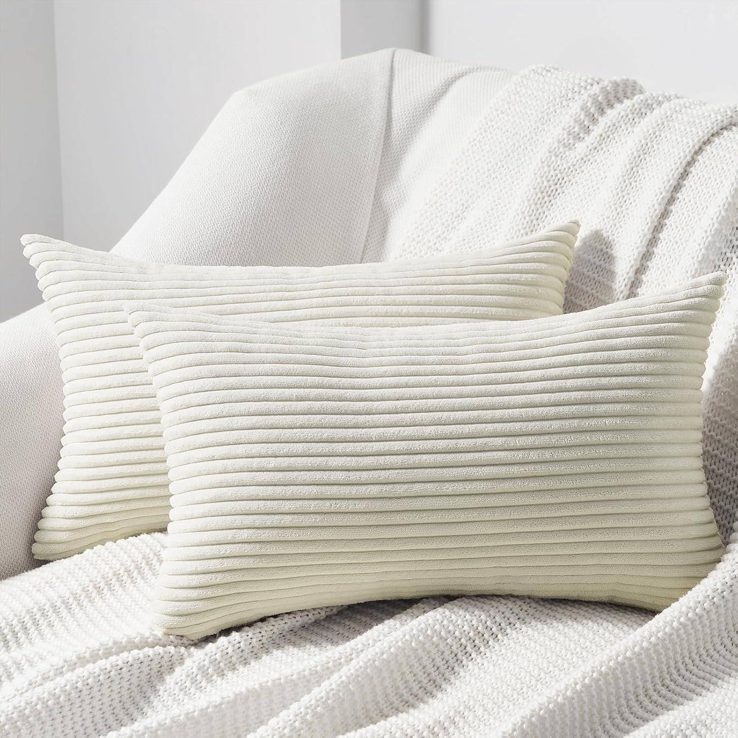 Zwei cremefarbene gerippte rechteckige Dekokissen auf einem weißen strukturierten Sessel mit einer gestrickten Decke, die eine weiche, neutrale Wohnzimmeratmosphäre schafft.