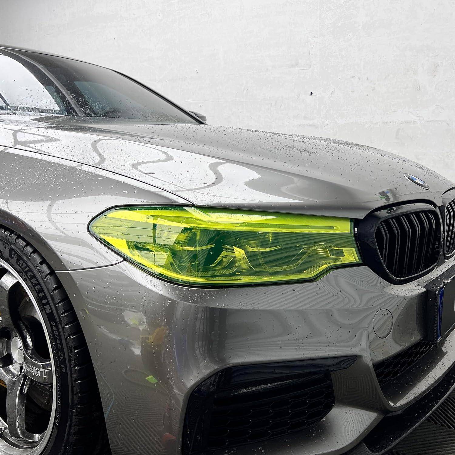 Nahaufnahme der vorderen linken Seite eines grauen BMW, die einen neongelb getönten Scheinwerfer, einen schwarzen Kühlergrill und eine nasse Karosserie zeigt.