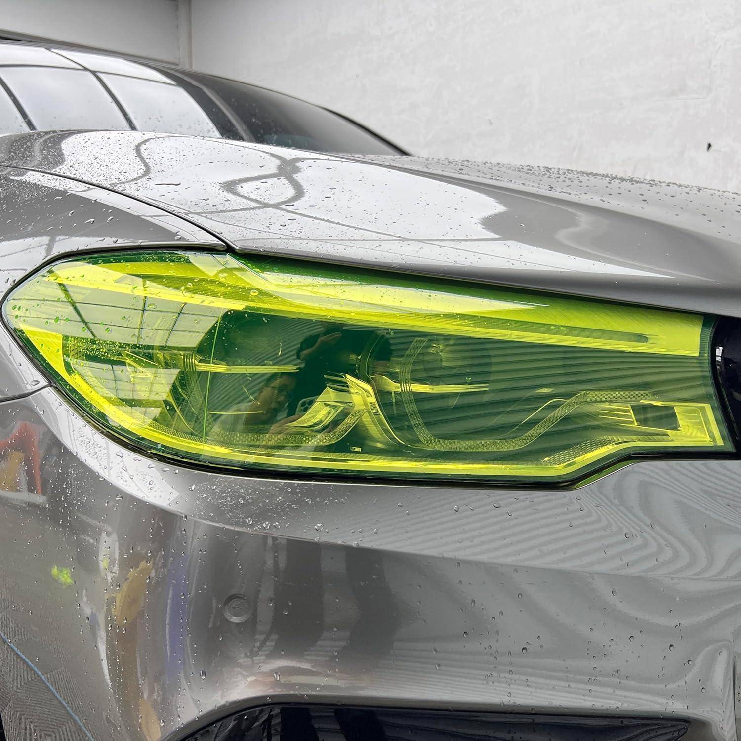 Nahaufnahme des vorderen Scheinwerfers eines silbernen Autos mit einer hell gelbgetönten Linse, nass von Regentropfen, vor einer einfachen Wand.