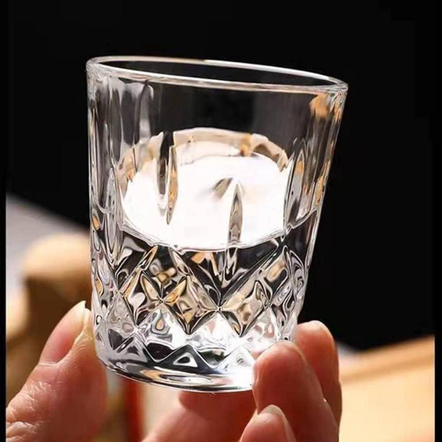 Nahaufnahme einer Hand, die ein kleines klares facettiertes Glas hält, das teilweise mit Wasser gefüllt ist, vor einem dunklen Hintergrund.