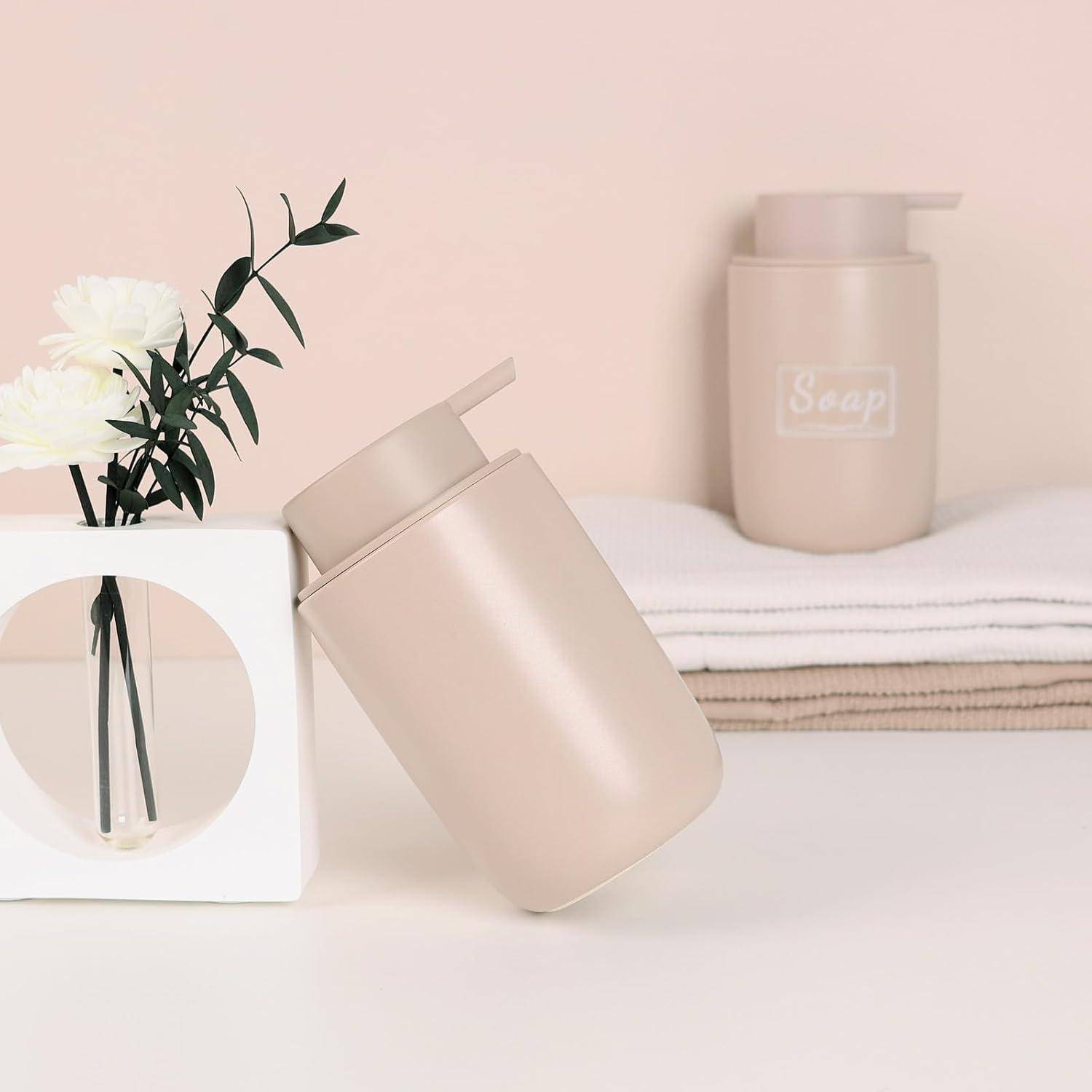 Zwei beige Seifenspender und ein weißes Blumenarrangement in einer minimalistischen Badezimmereinrichtung, wobei eine Pumpflasche gegen einen weißen Rahmen lehnt.