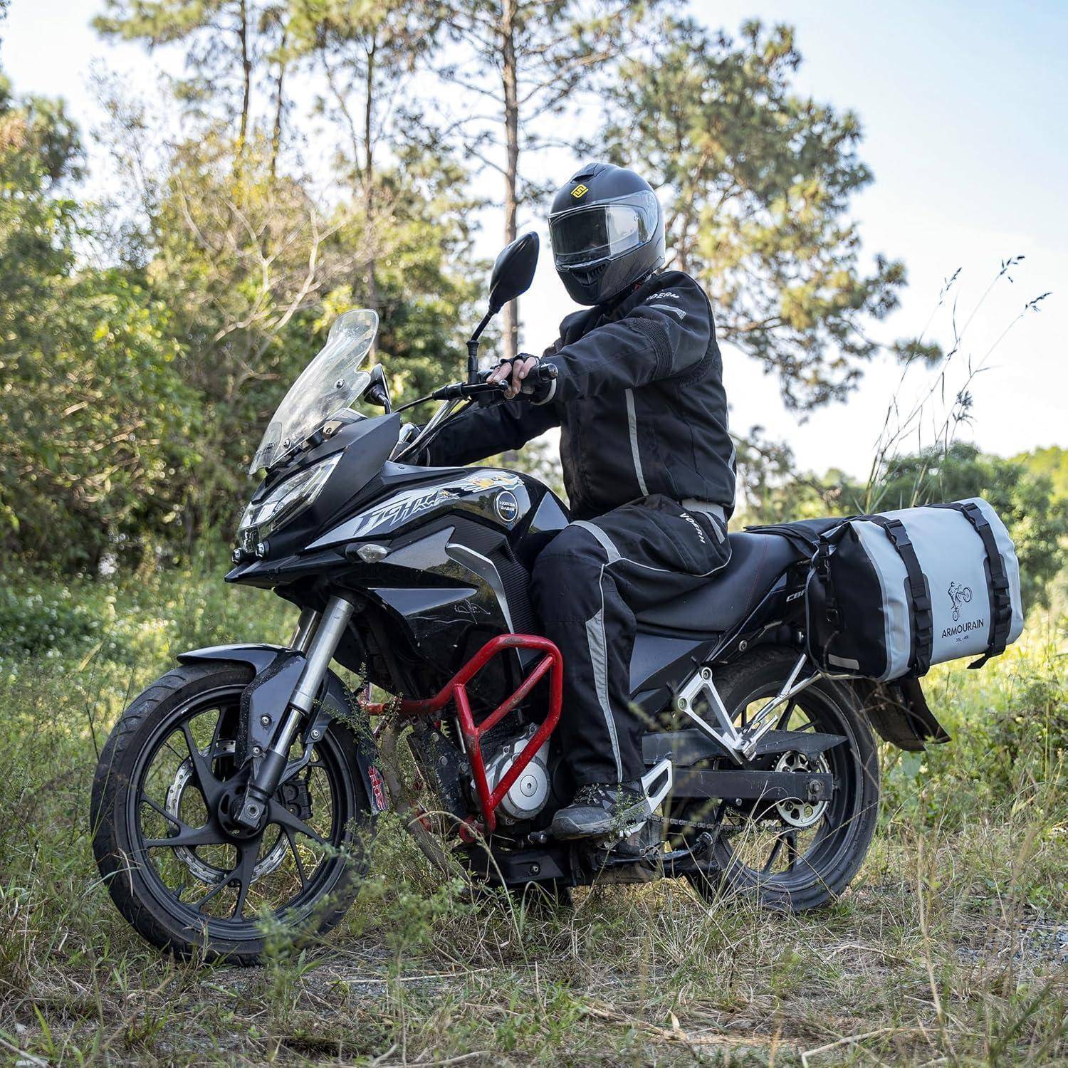 Ein Motorradfahrer mit Helm sitzt auf einem schwarzen Adventure-Motorrad mit beladenen Seitentaschen, das auf grasbewachsenem Grund in einem Waldgebiet geparkt ist.