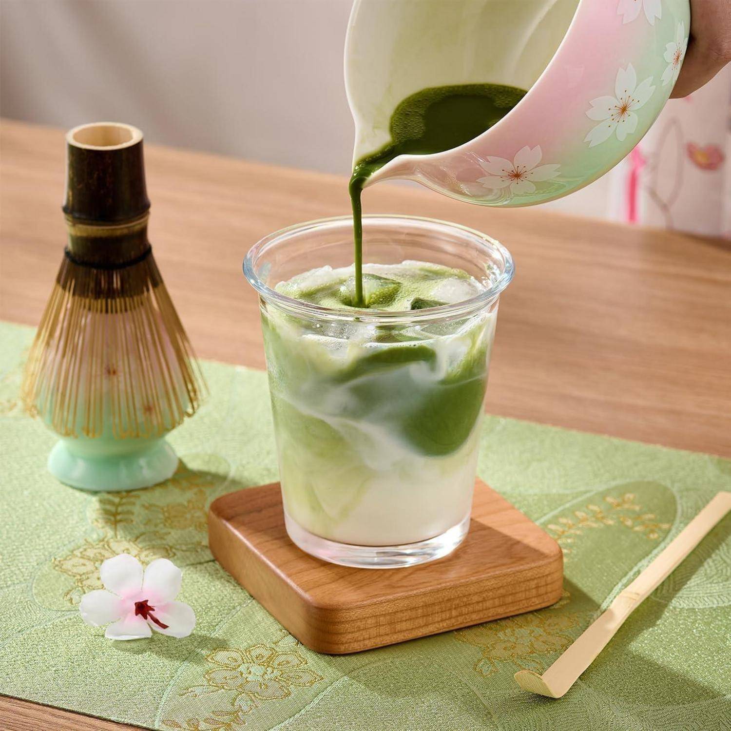 Grüner Matcha-Tee wird in ein Glas mit Milch auf einem Holzuntersetzer gegossen, mit einem Bambusbesen und einem Löffel auf einer Teematte.