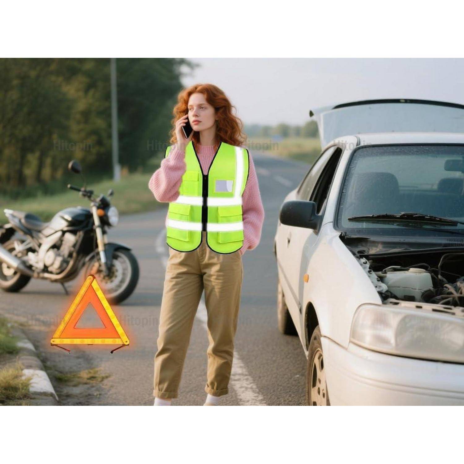 Frau in einer reflektierenden Weste, die neben einem liegen gebliebenen weißen Auto mit offener Motorhaube telefoniert, mit einem Warndreieck am Straßenrand und einem Motorrad im Hintergrund.