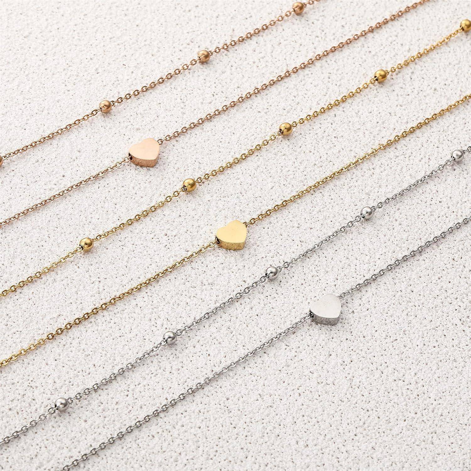Fünf zarte Kettenarmbänder in Gold, Roségold und Silber, jedes mit kleinen Perlenakzenten und herzförmigen Anhängern auf einem hell strukturierten Hintergrund.