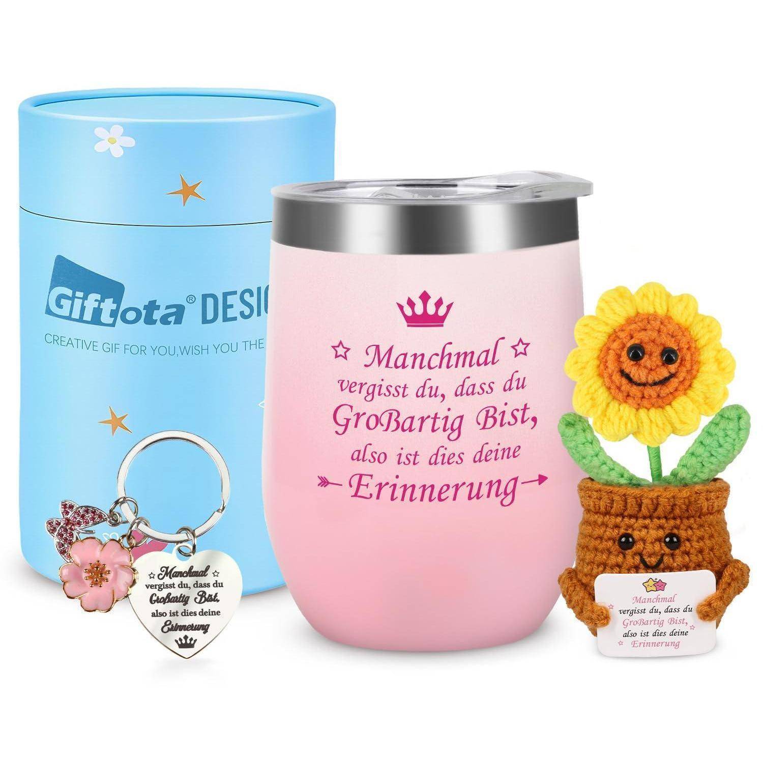 Geschenkset mit isoliertem rosa Tumbler, blauer Geschenkbox, Herz-Schlüsselanhänger und gehäkelter lächelnder Blume im Topf; deutscher Text auf Tumbler und Topf.