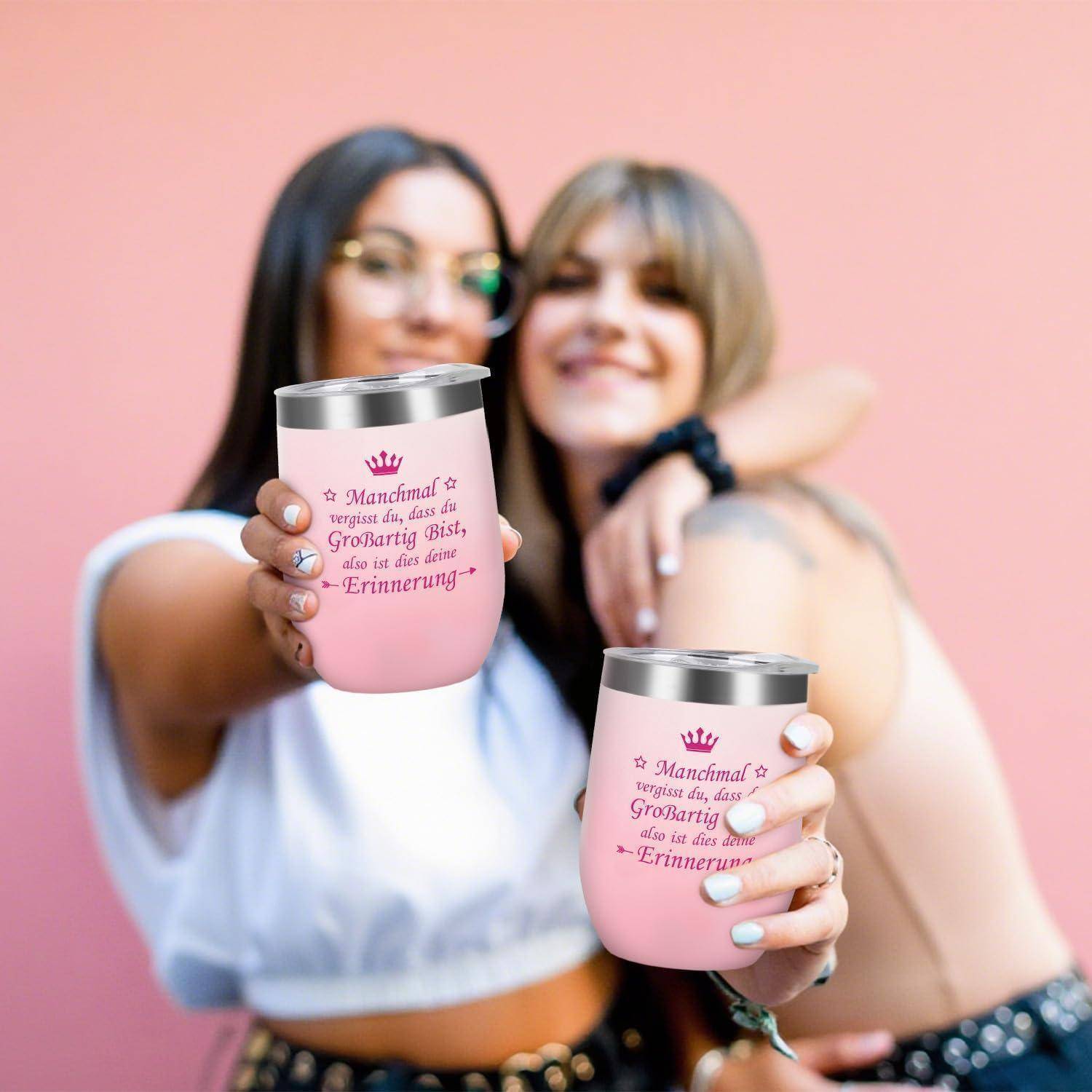 Zwei Frauen lächeln und halten passende rosa Trinkbecher mit deutscher Aufschrift in Richtung der Kamera vor einem rosa Hintergrund.