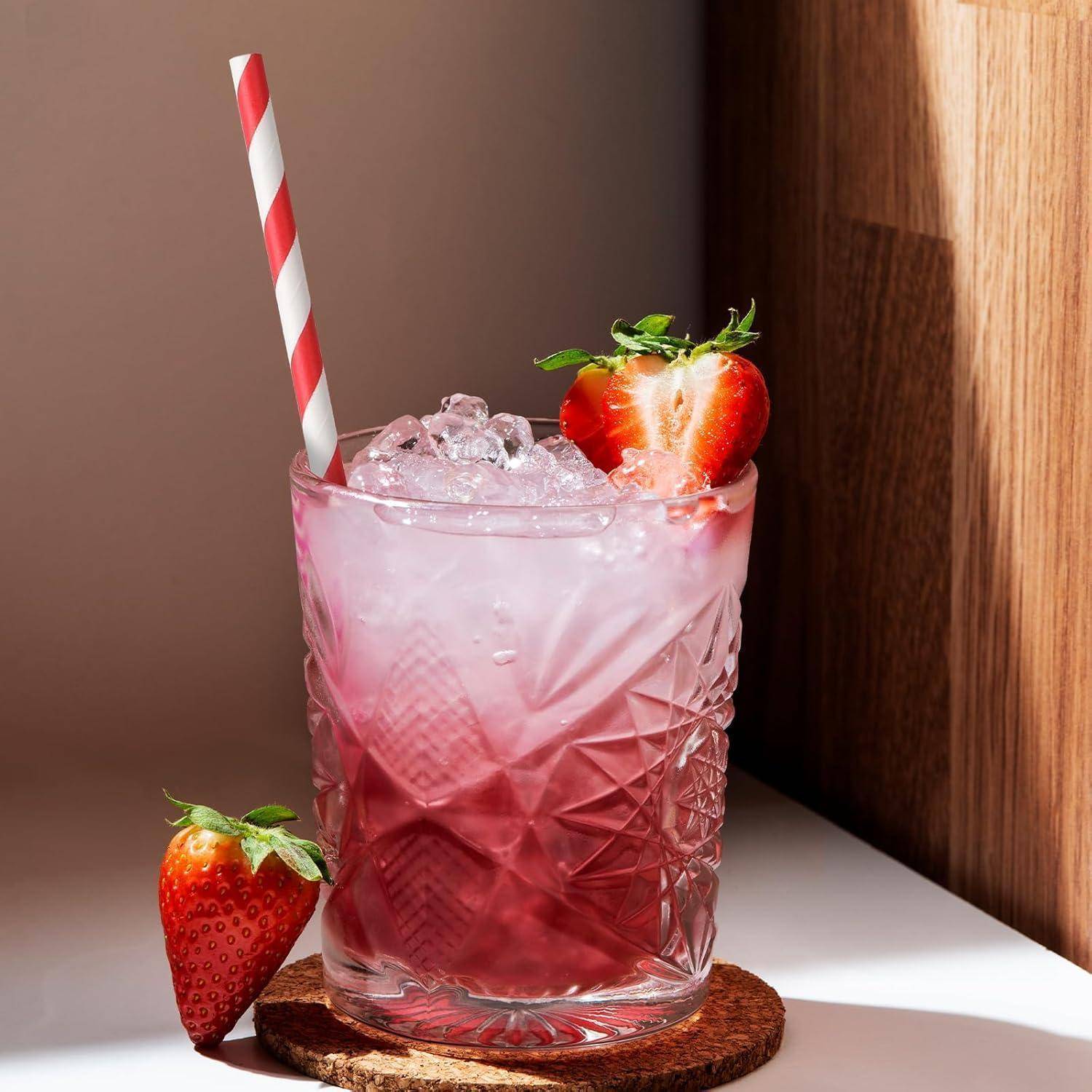 Rosiger Erdbeer-Cocktail in einem facettierten Glas mit Eis, einem gestreiften Strohhalm und frischen Erdbeeren, stehend auf einem Untersetzer neben einer Holzwand.