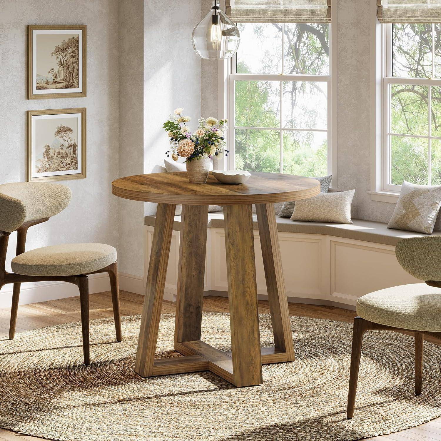 Ein runder Holztisch mit einer Blumengesteck-Mitteldekoration steht in einer hellen Essecke mit zwei gepolsterten Stühlen, einer Fensterbank und gerahmten Wandkunst.