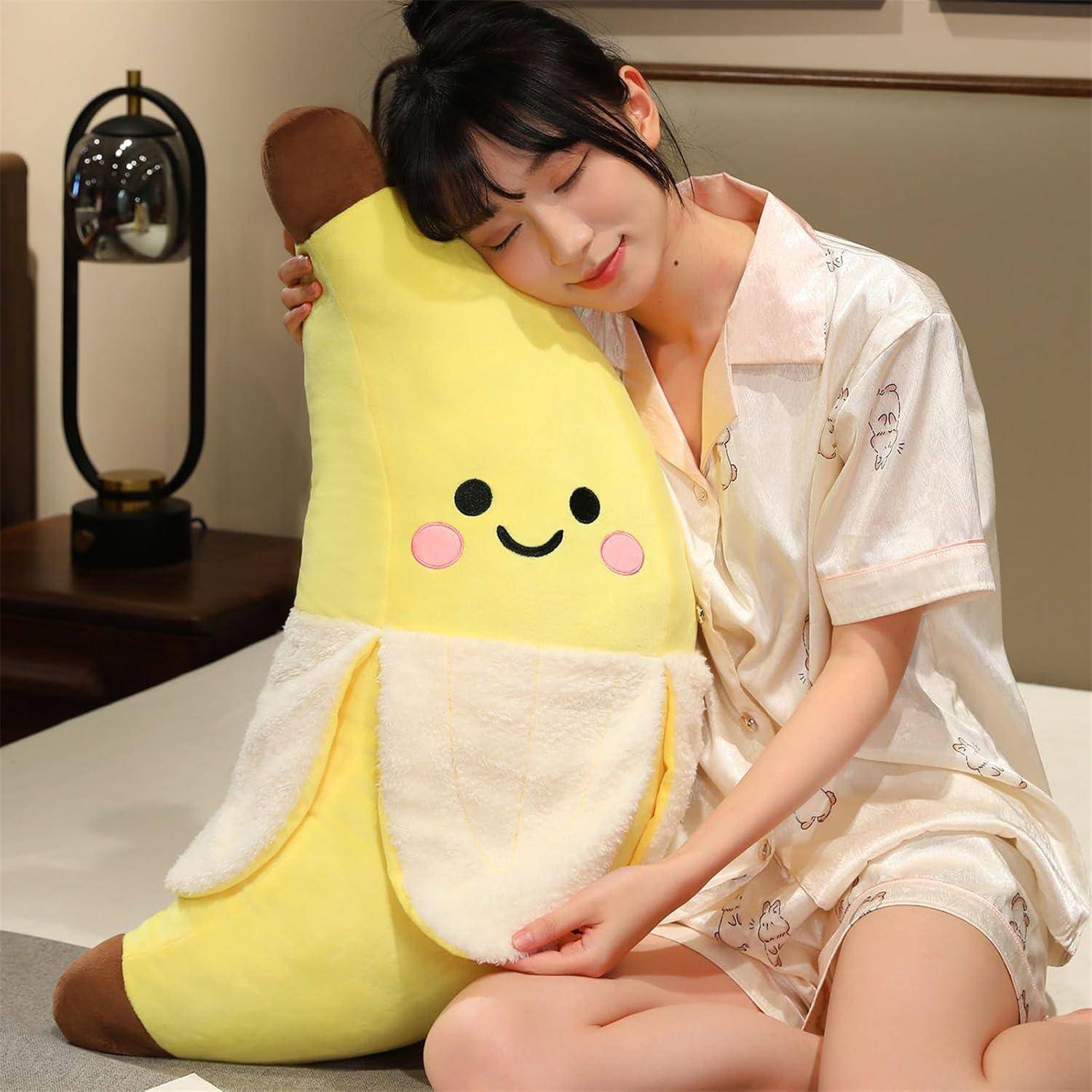 Junge Frau in heller Pyjama-Kleidung, die ein großes lächelndes bananenförmiges Plüschtier umarmt, während sie auf einem Bett sitzt.