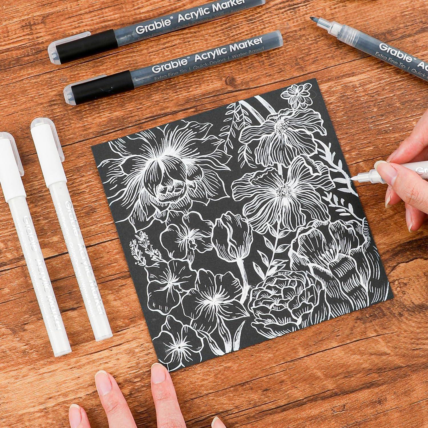 Handgezeichnete weiße florale Strichkunst auf einer schwarzen quadratischen Oberfläche mit Acrylmarkern auf einem Holztisch.