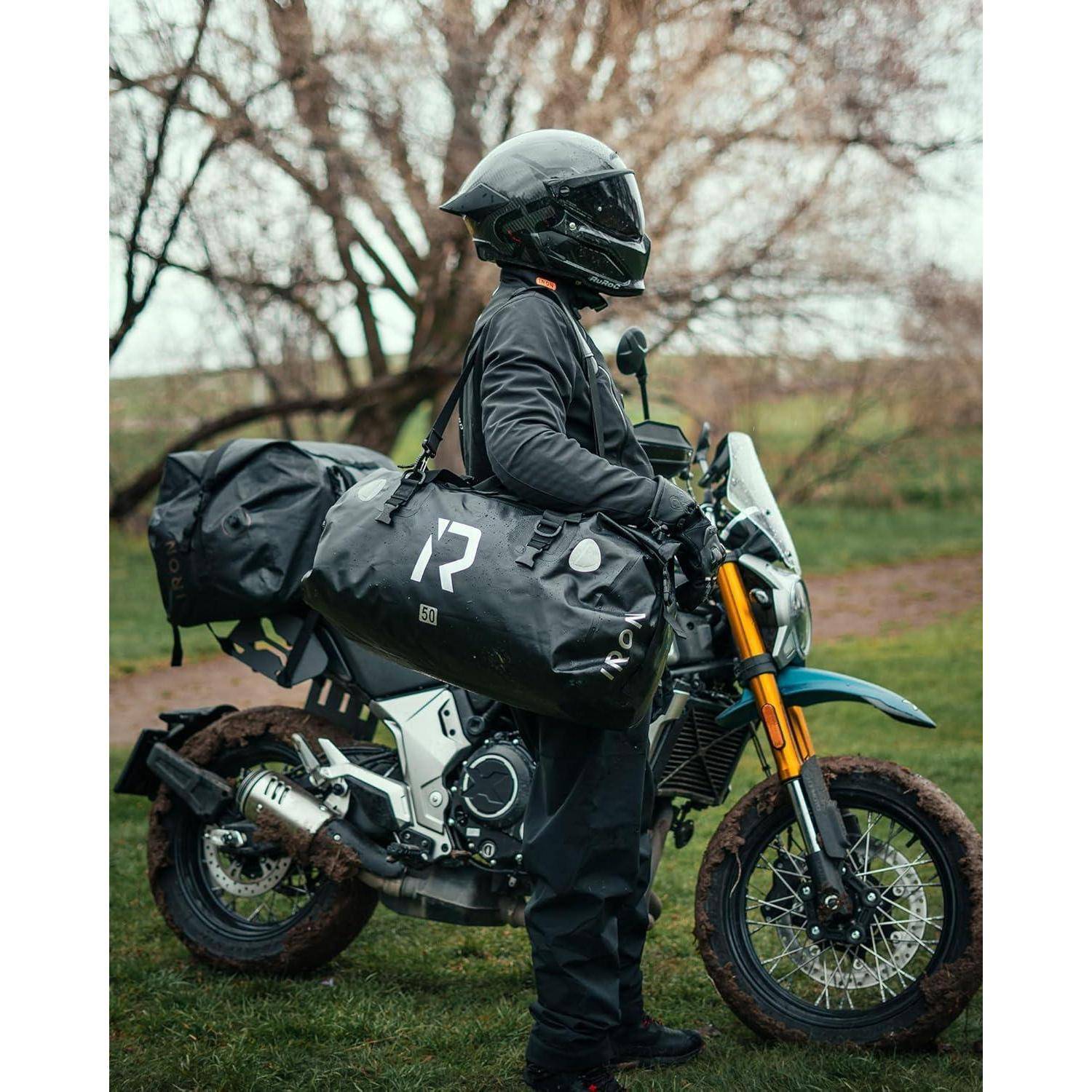Motorradfahrer in schwarzer Ausrüstung und Helm sitzt auf einem Abenteuermotorrad, das mit Seitentaschen beladen ist, geparkt auf schlammigem Gras in einer ländlichen Umgebung.