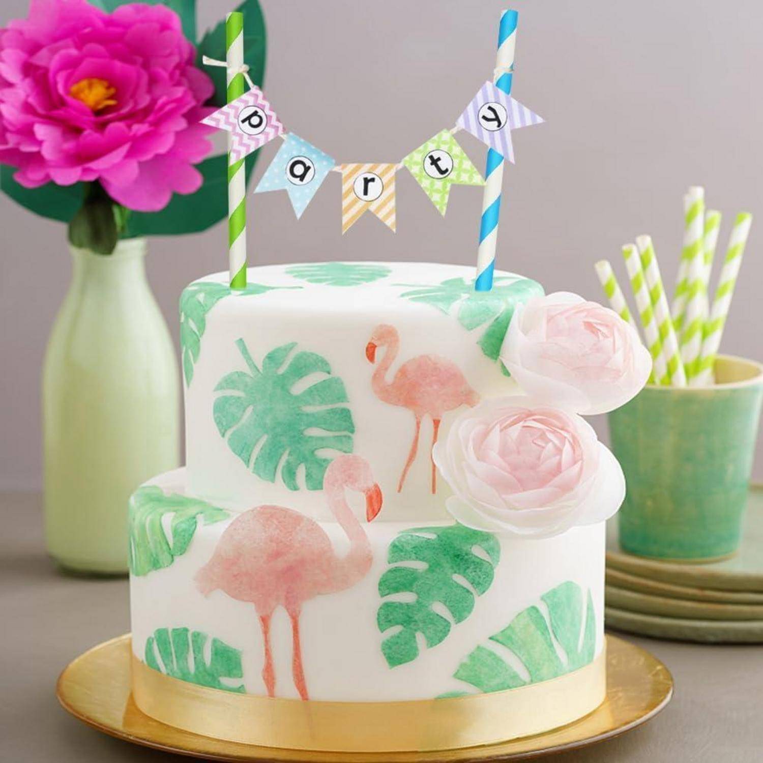 Zweistöckige weiße Geburtstagstorte mit Flamingo- und tropischen Blattdekorationen, rosa Rosen und "Party""-Wimpeln auf Tortenspitzern.