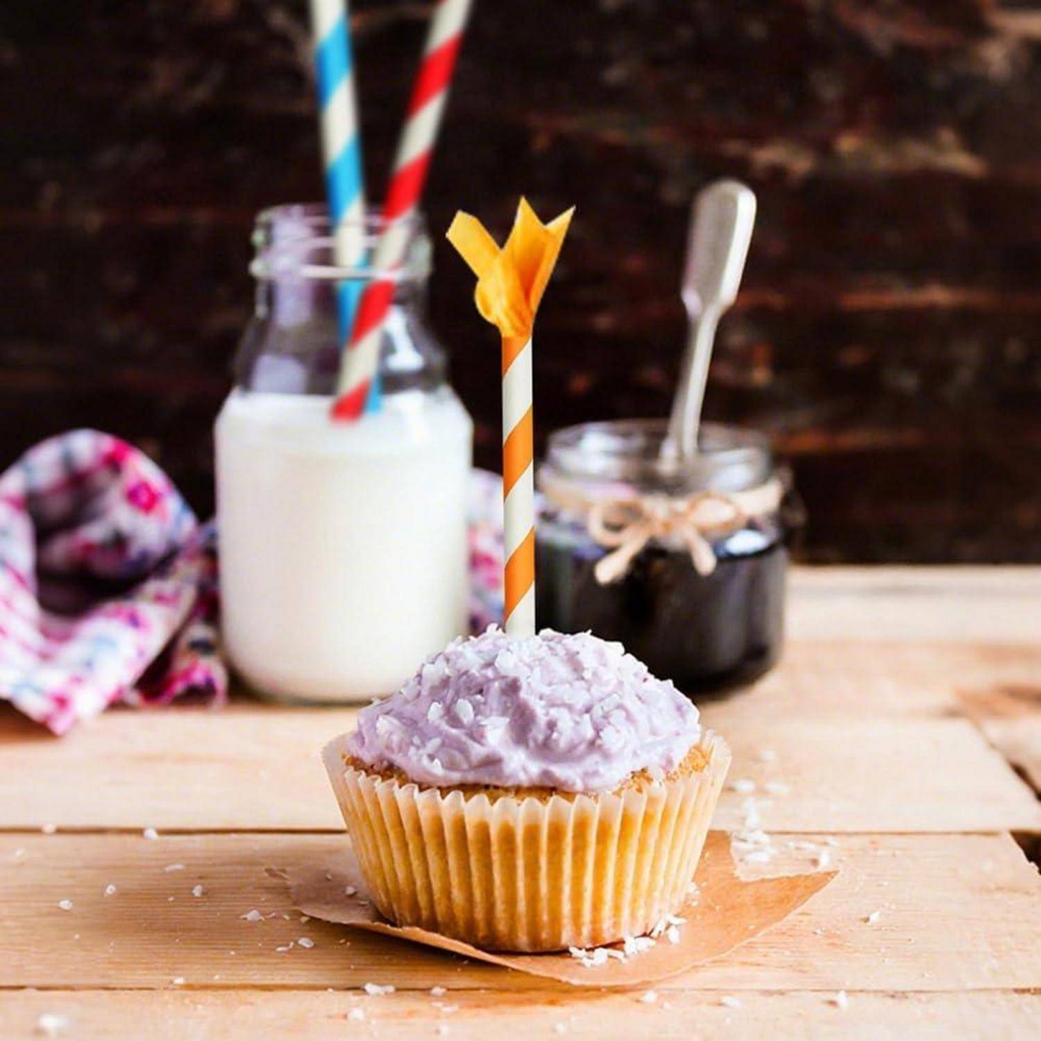 Ein gefrosteter Cupcake mit violettem Frosting und einer orange gestreiften Kerze auf einem Holzbrett, mit Milch und einem Glas im Hintergrund.