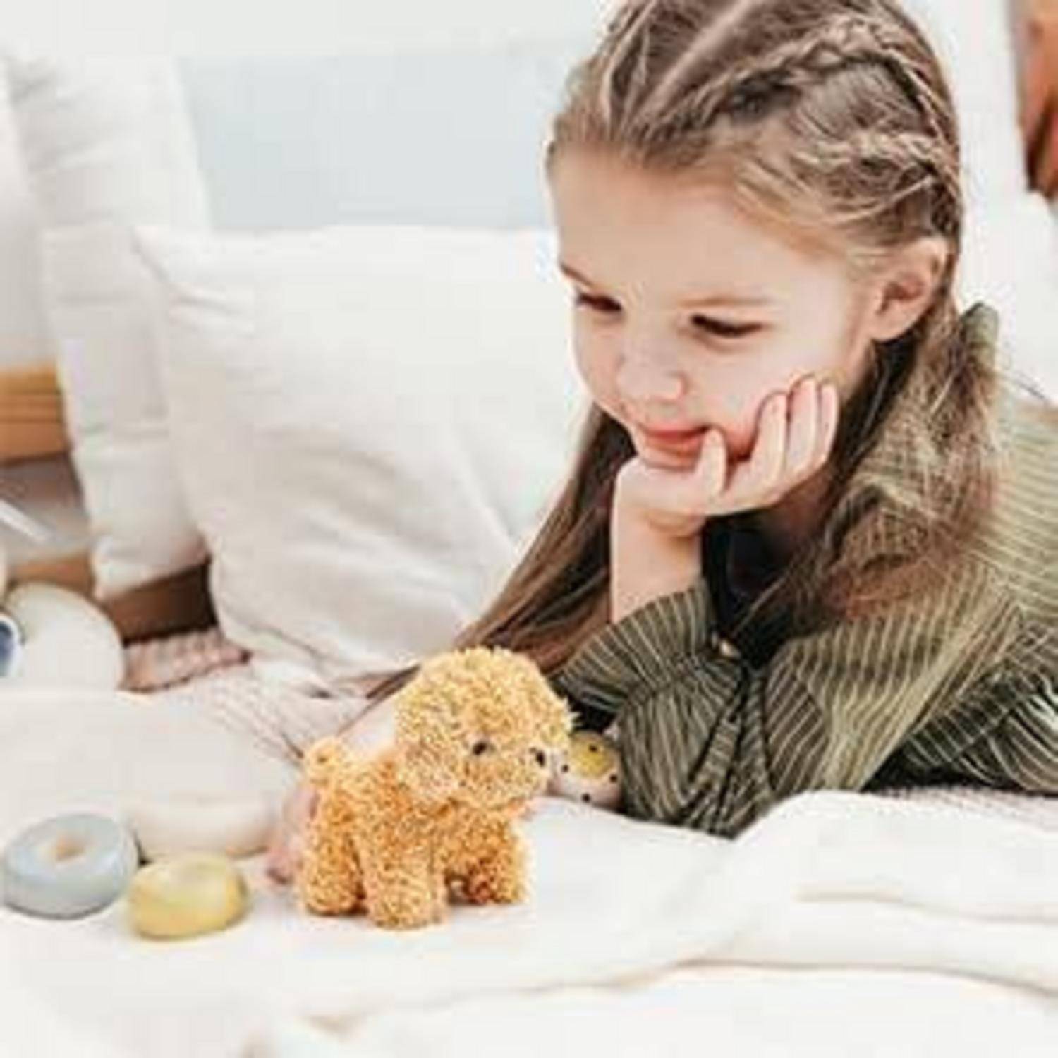 Junges Mädchen, das auf einem Bett liegt, sein Kinn auf der Hand abstützt und auf ein kleines Teddybär-Spielzeug in der Nähe schaut.