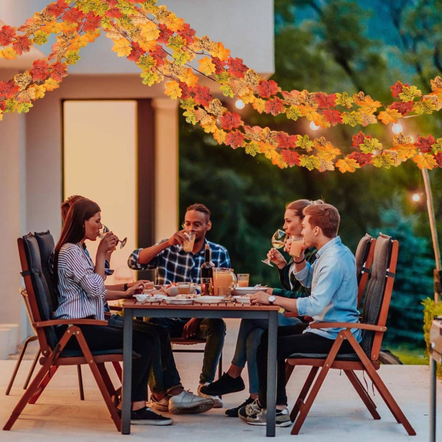 Vier Freunde sitzen an einem Außentisch und essen und trinken zusammen unter Herbstlaubgirlanden auf einer Terrasse in der Dämmerung.