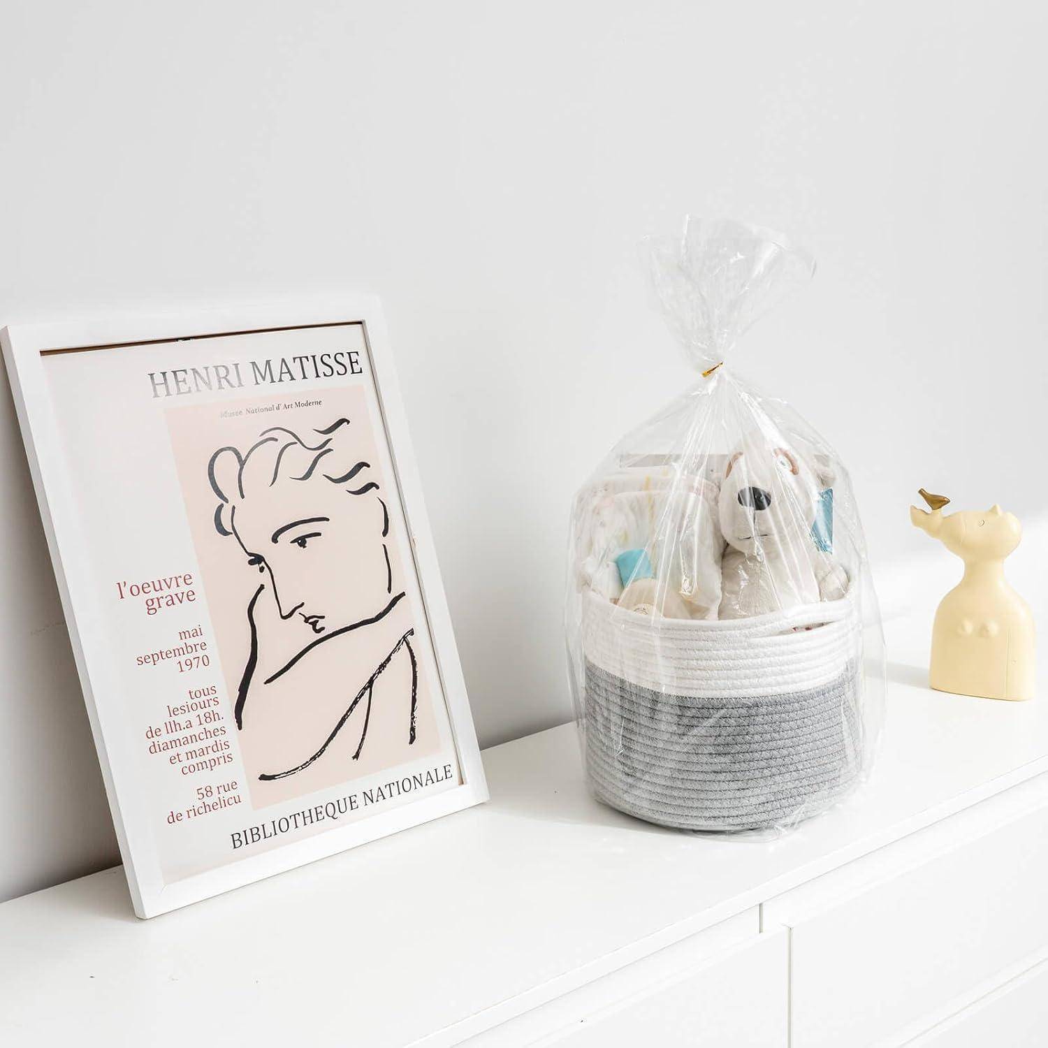 Gerahmtes Henri-Matisse-Plakat auf einer weißen Kommode neben einem verpackten Geschenkkorb mit einem Teddybär und einer kleinen gelben Dekorationsfigur.