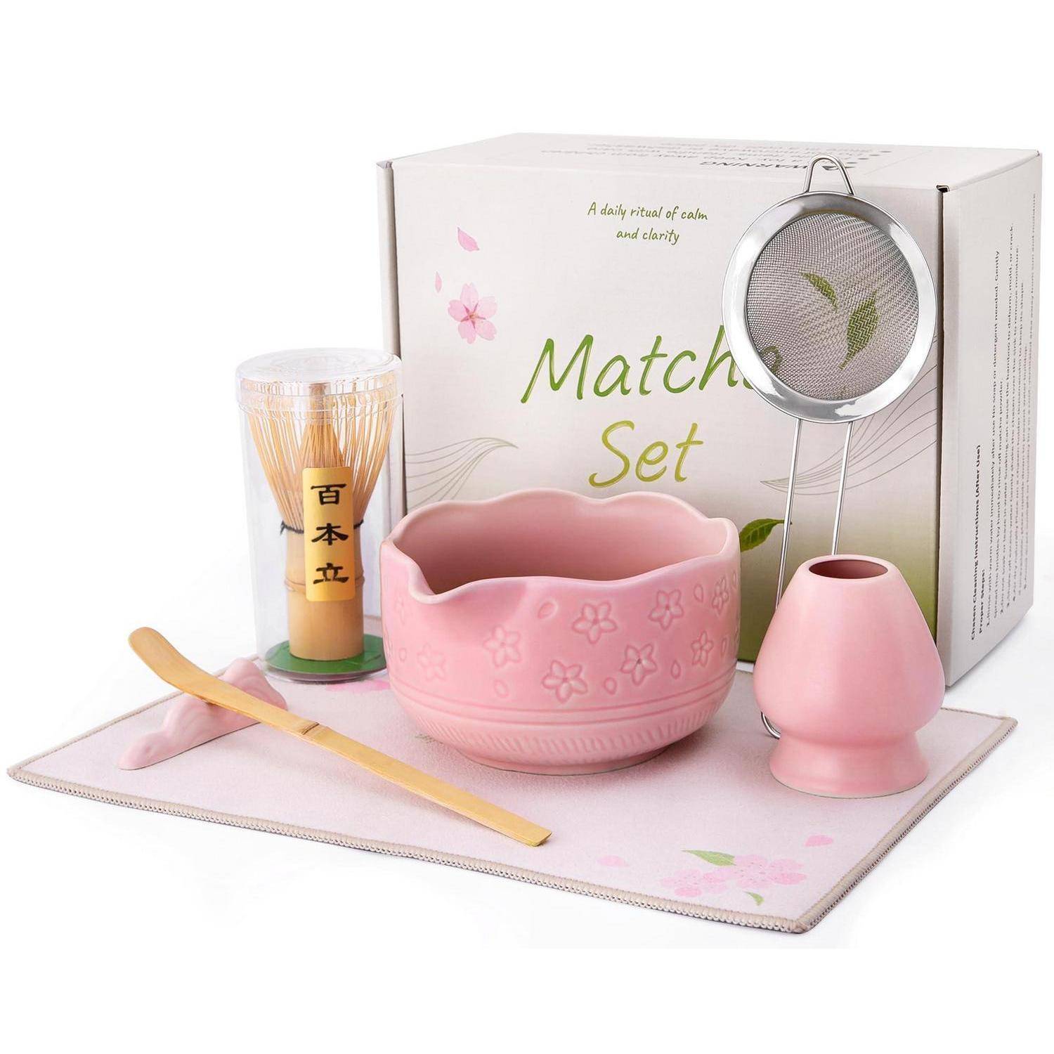 Rosa Matcha-Tee-Set mit Schüssel, Besen, Löffel, Sieb, Tasse und Schachtel auf einer Matte.