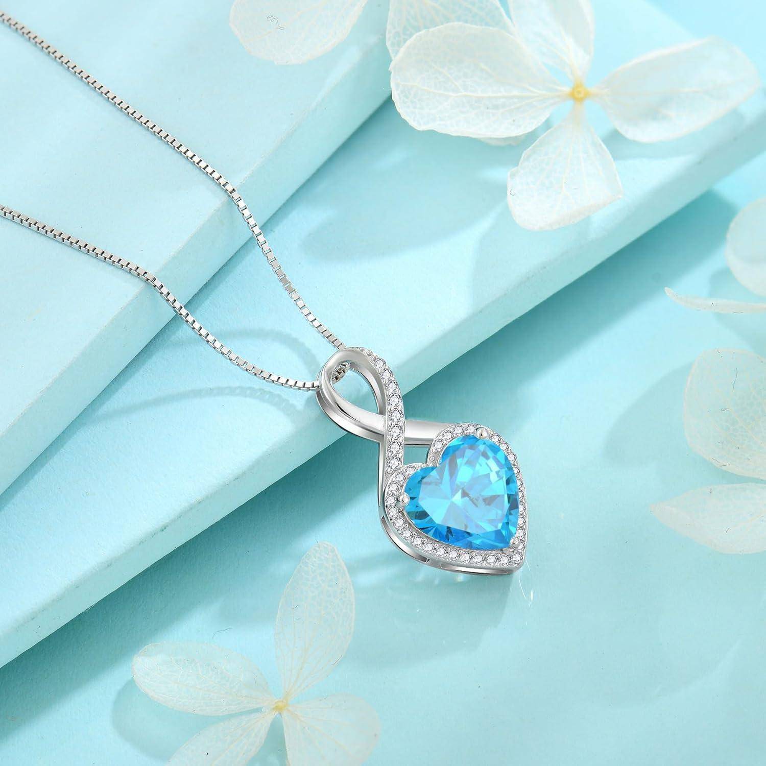 Silberkette mit anhängendem blauem herzförmigem Anhänger, der mit transparenten Steinen umrandet ist, präsentiert auf einer hellblauen Geschenkbox mit weißen Blumen.