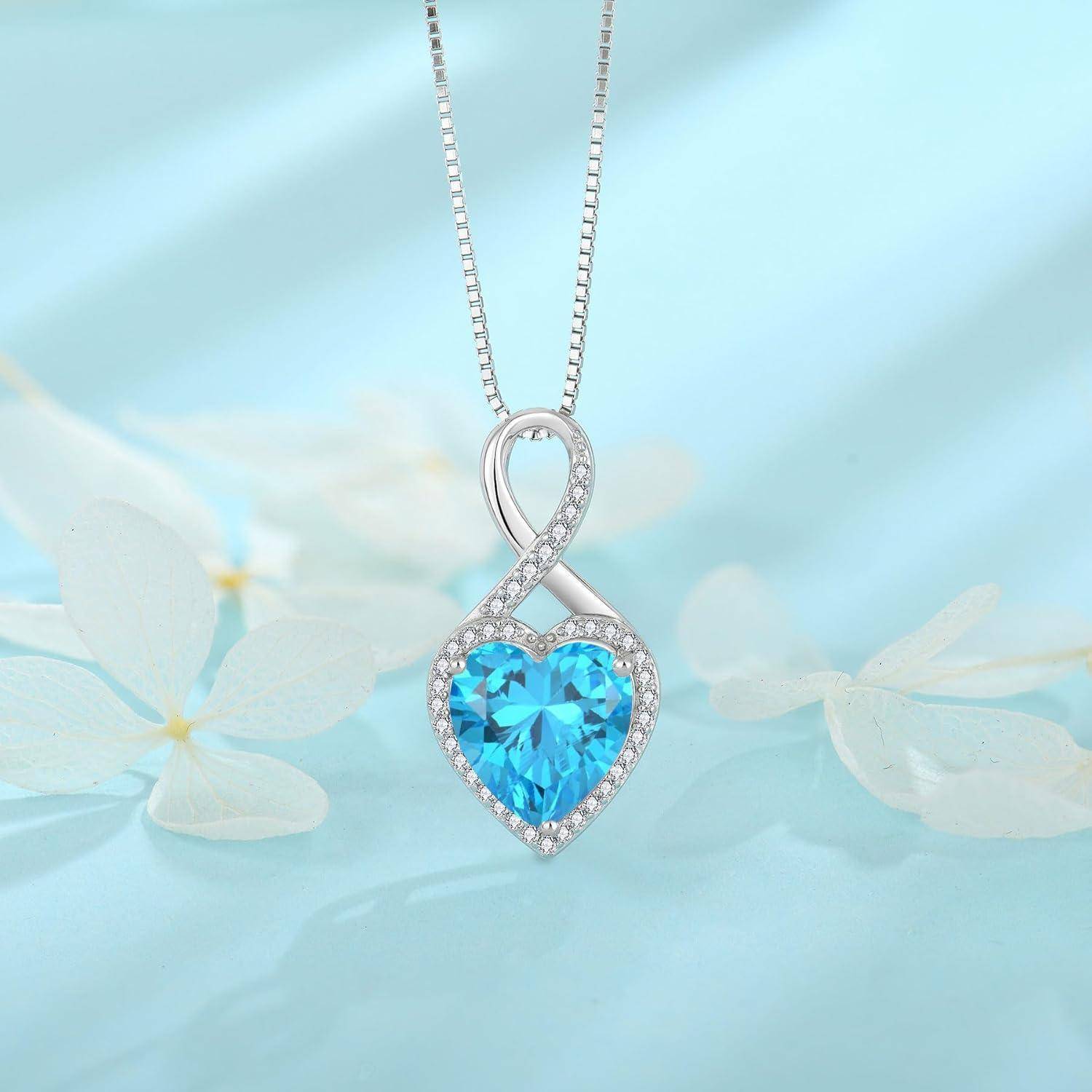Eine silberne Unendlichkeits-Herz-Anhänger-Halskette mit einem leuchtend blauen herzförmigen Edelstein und kleinen klaren Steinen, gezeigt vor einem sanften blauen Blumenhintergrund.